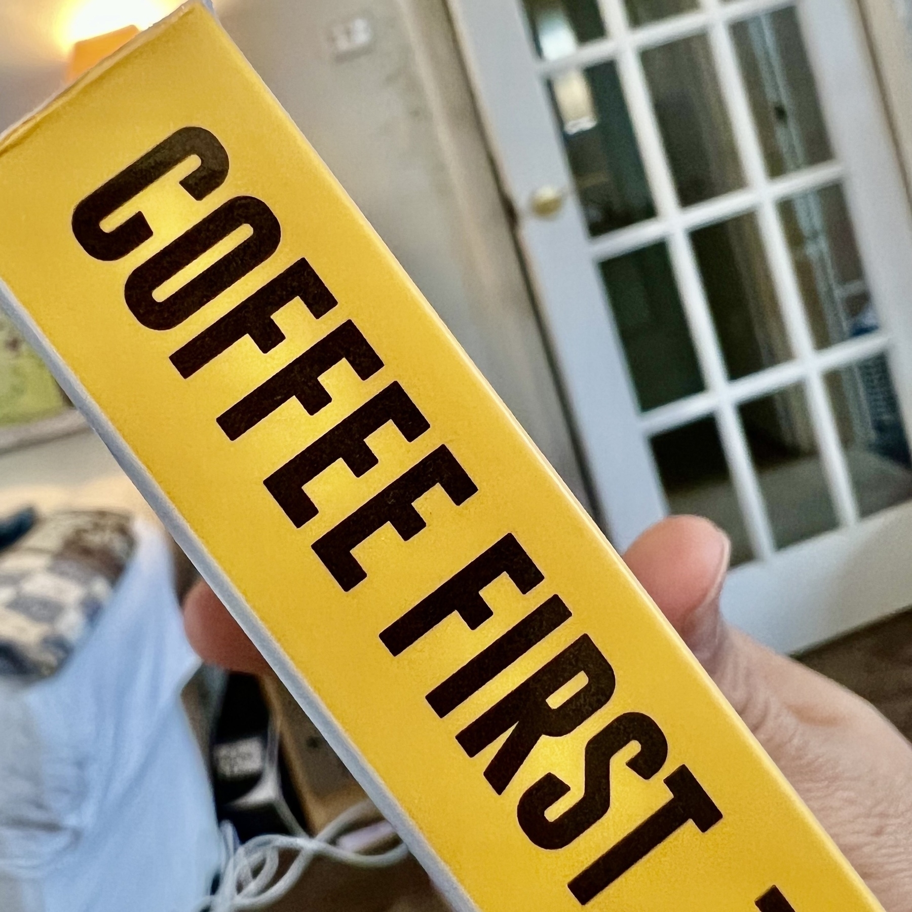 A person holds a book with the yellow spine titled "COFFEE FIRST" in a room with a sofa and a glass-paned door in the background.