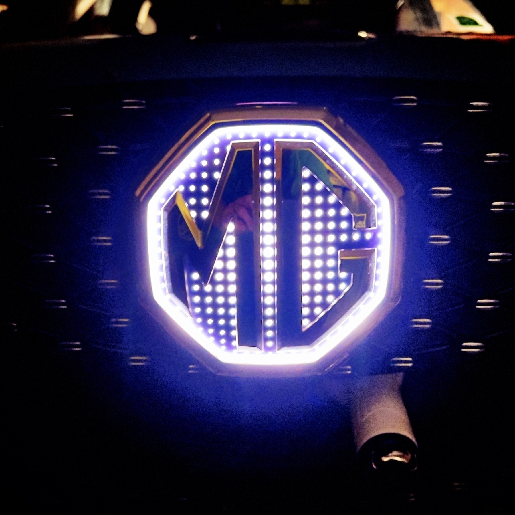 An illuminated MG emblem is prominently displayed on the front of a charging EV, surrounded by a glowing light.