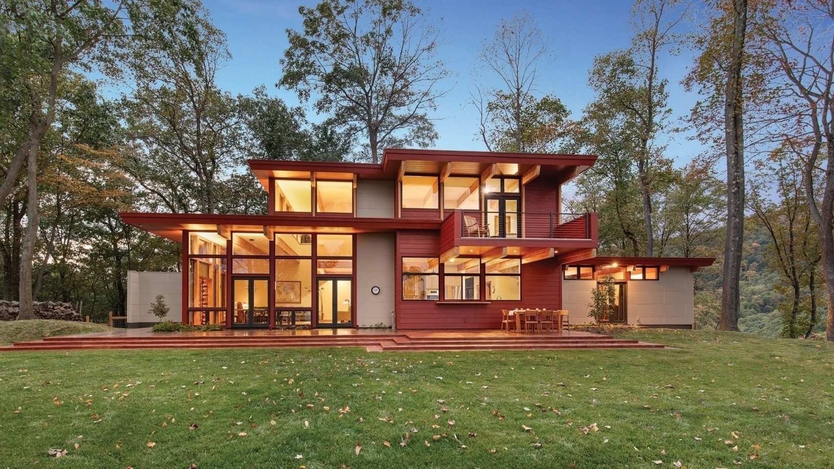 A modern, two-story house with large windows is situated in a lush, wooded area.