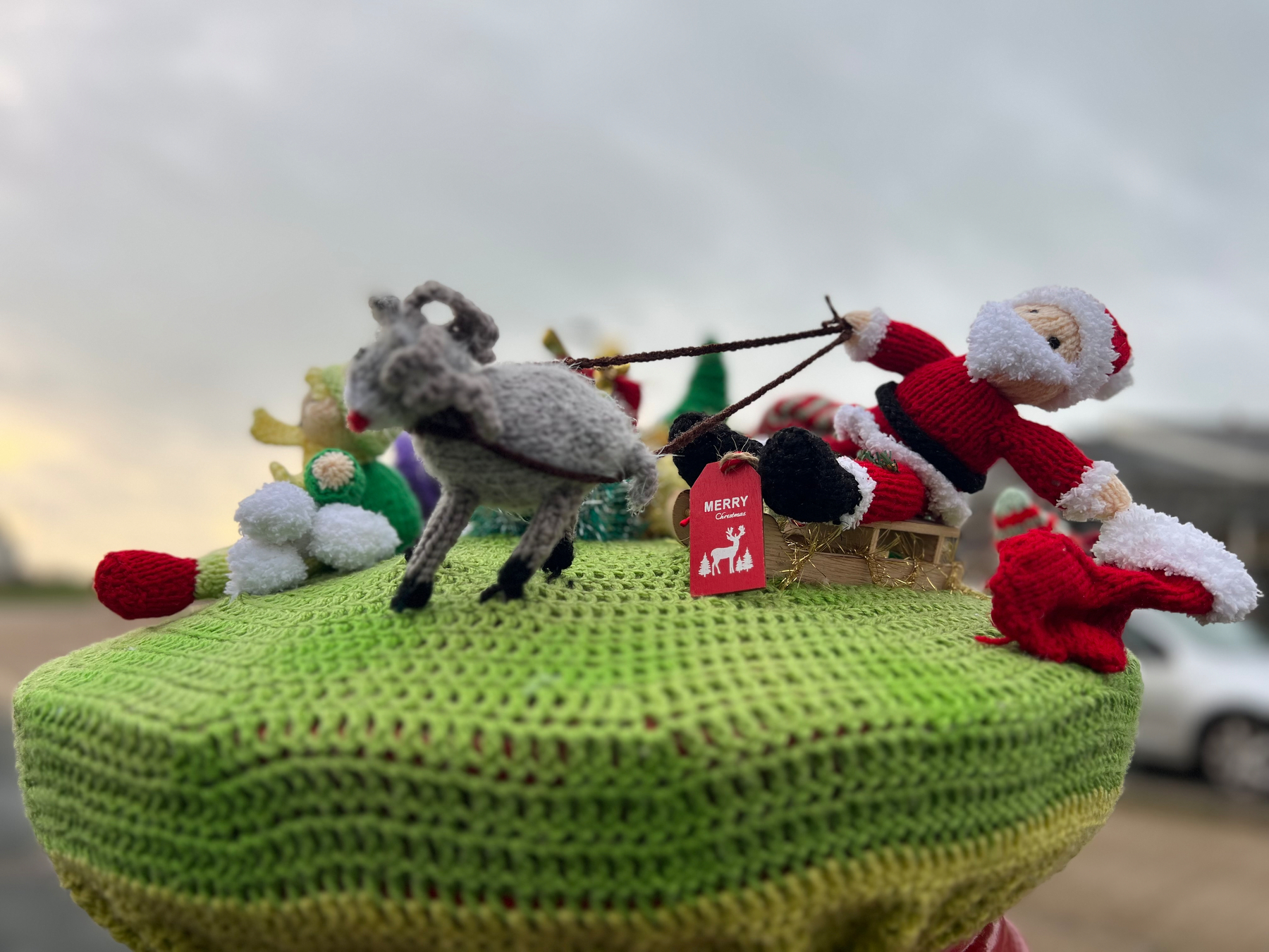 A knitted Santa Claus is depicted riding a sleigh pulled by a reindeer, sitting atop a green knitted surface with a Merry tag.