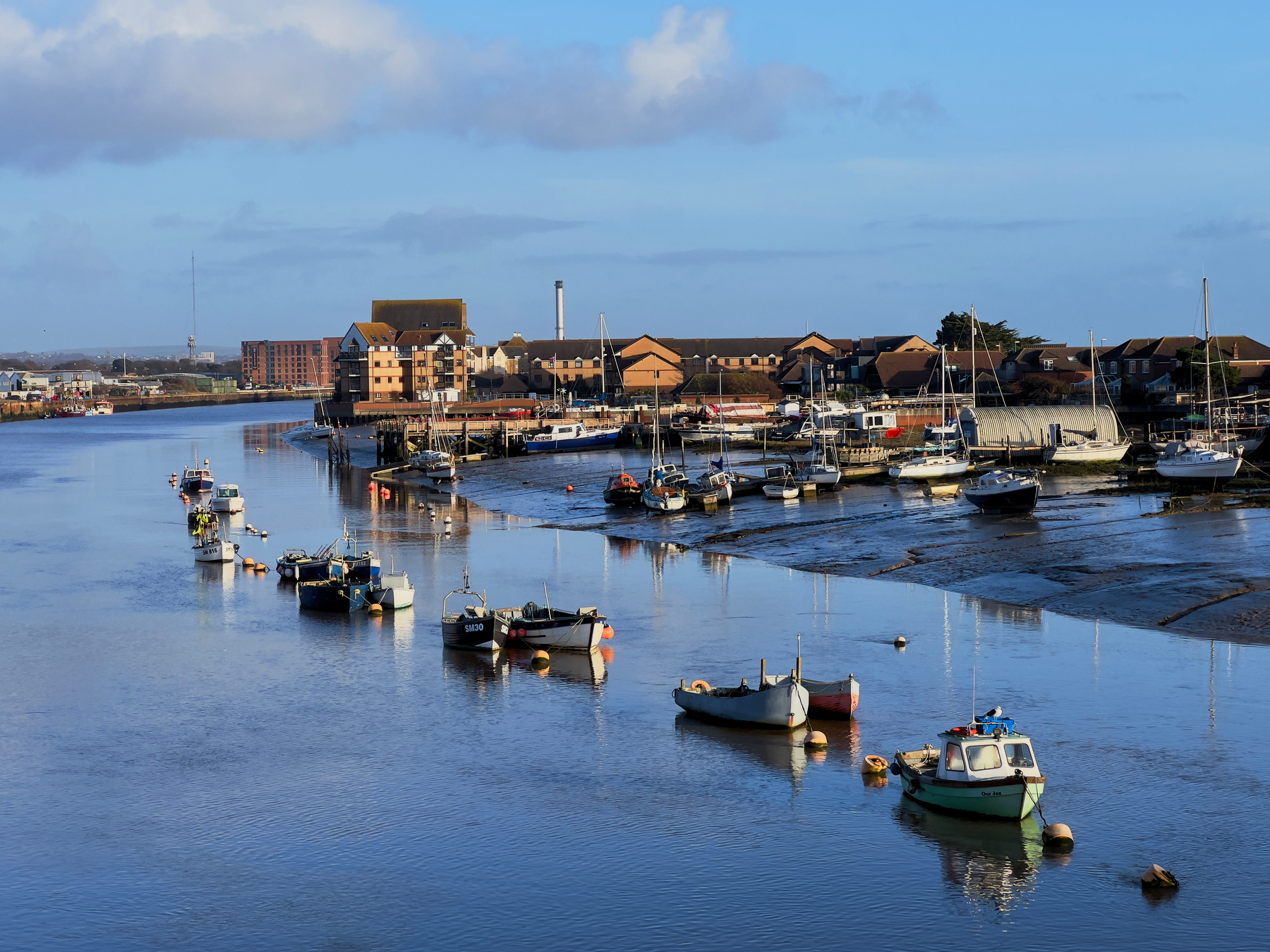 Small boats are anchored along a calm riverbank near a coastal town with buildings in the background.
