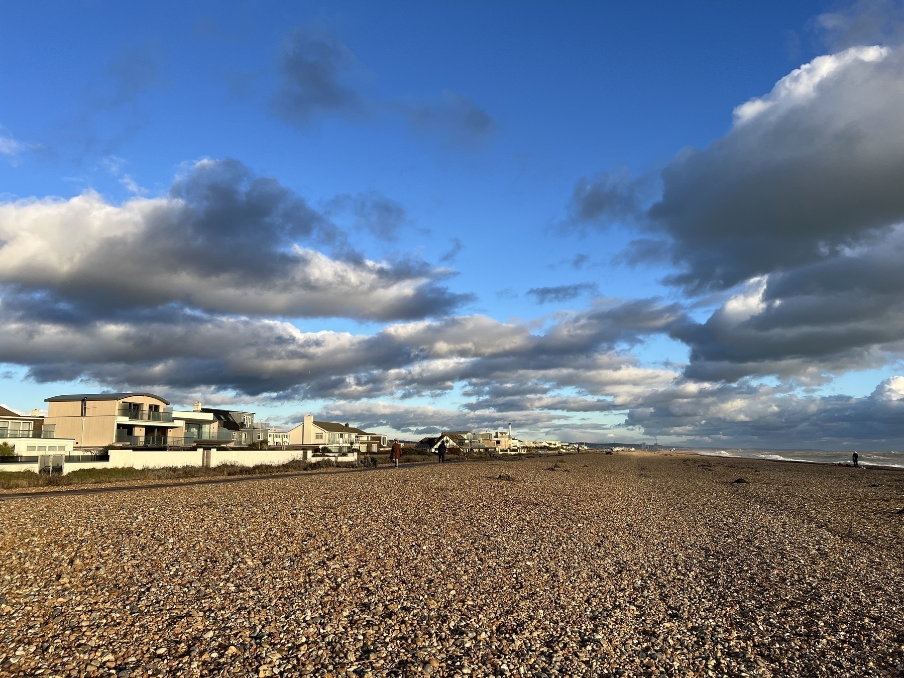 A pebble beach stretches along the coast under a partly cloudy sky, with modern houses lining the shore.