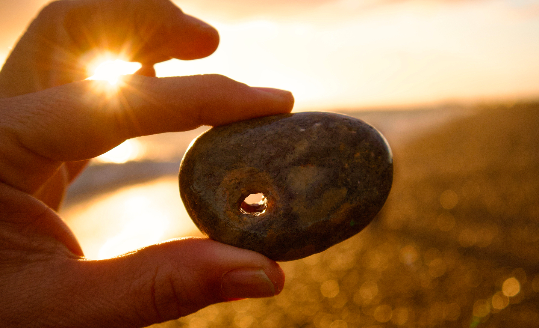 A hand holds a smooth stone with a hole in it against the backdrop of a sunlit beach.