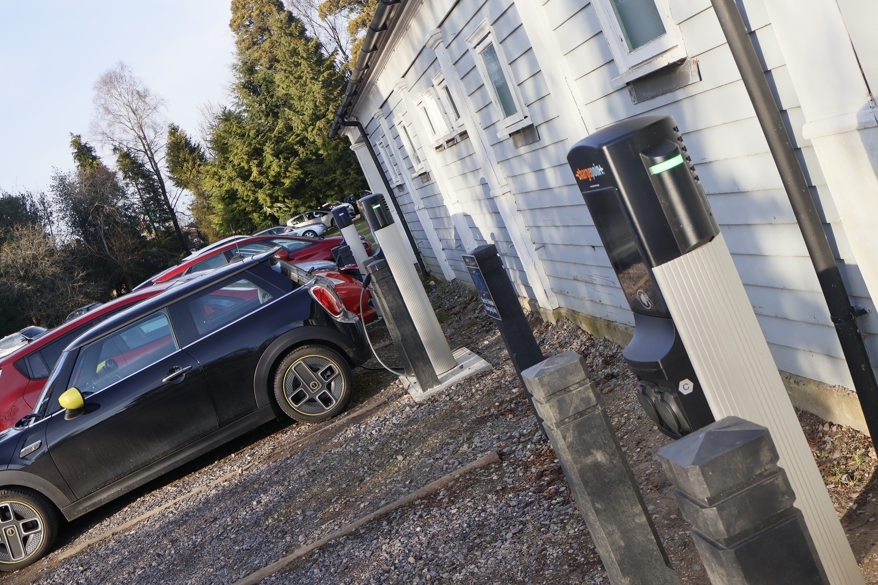 A row of electric cars is parked at a charging station beside a white building.