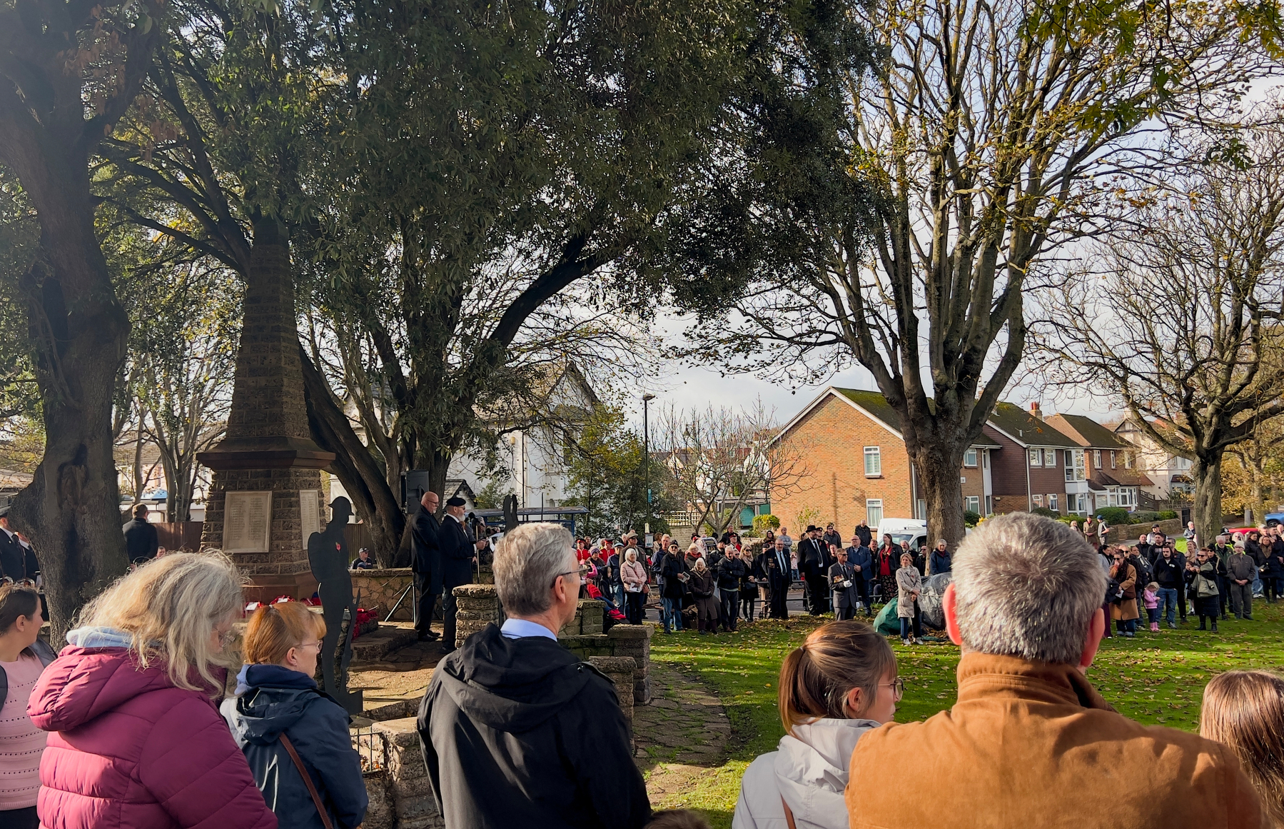 A group of people gathers in a park area near a stone monument, surrounded by trees and houses.