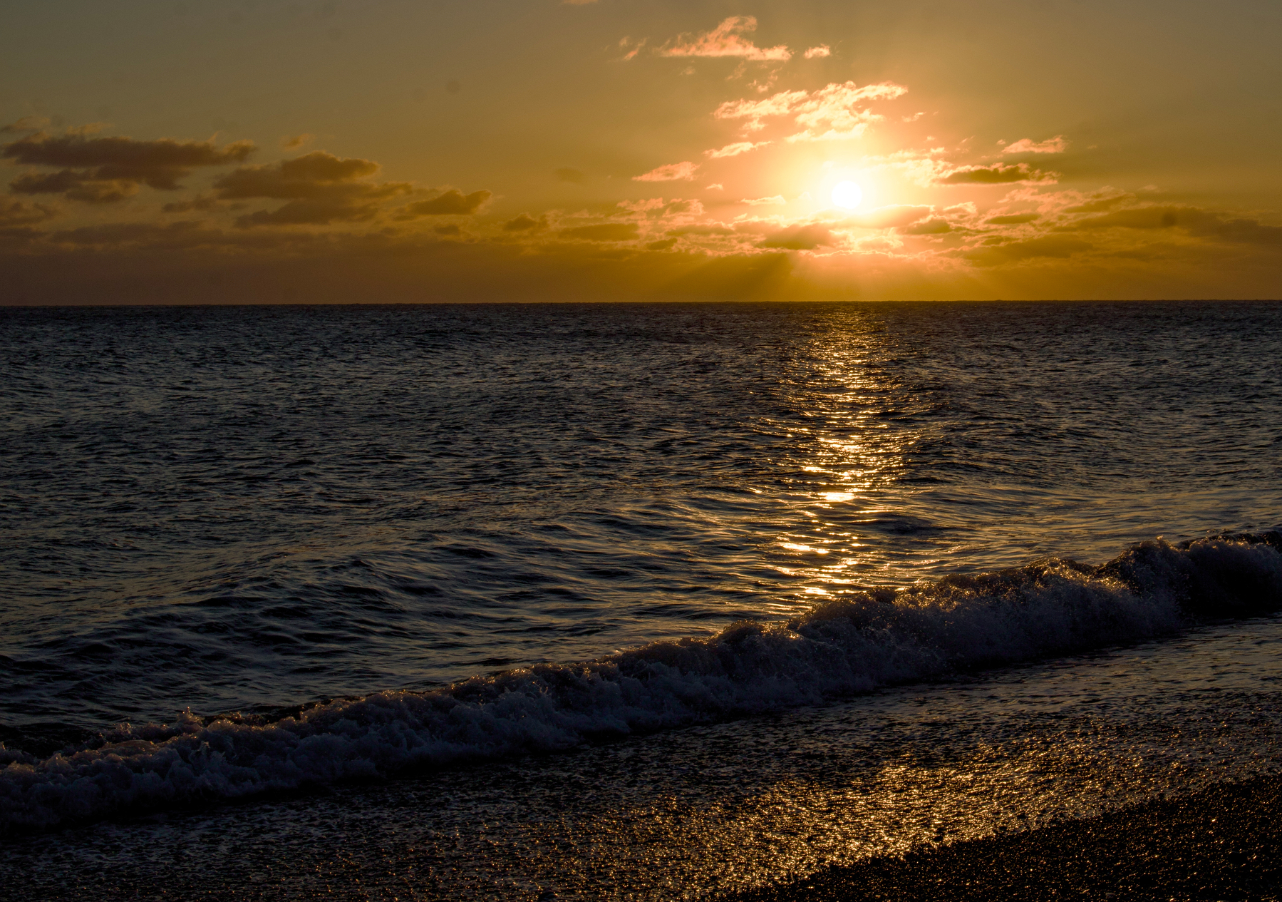 A serene beach scene features the sun setting over the ocean, casting a golden reflection on the water and waves gently washing onto the shore.