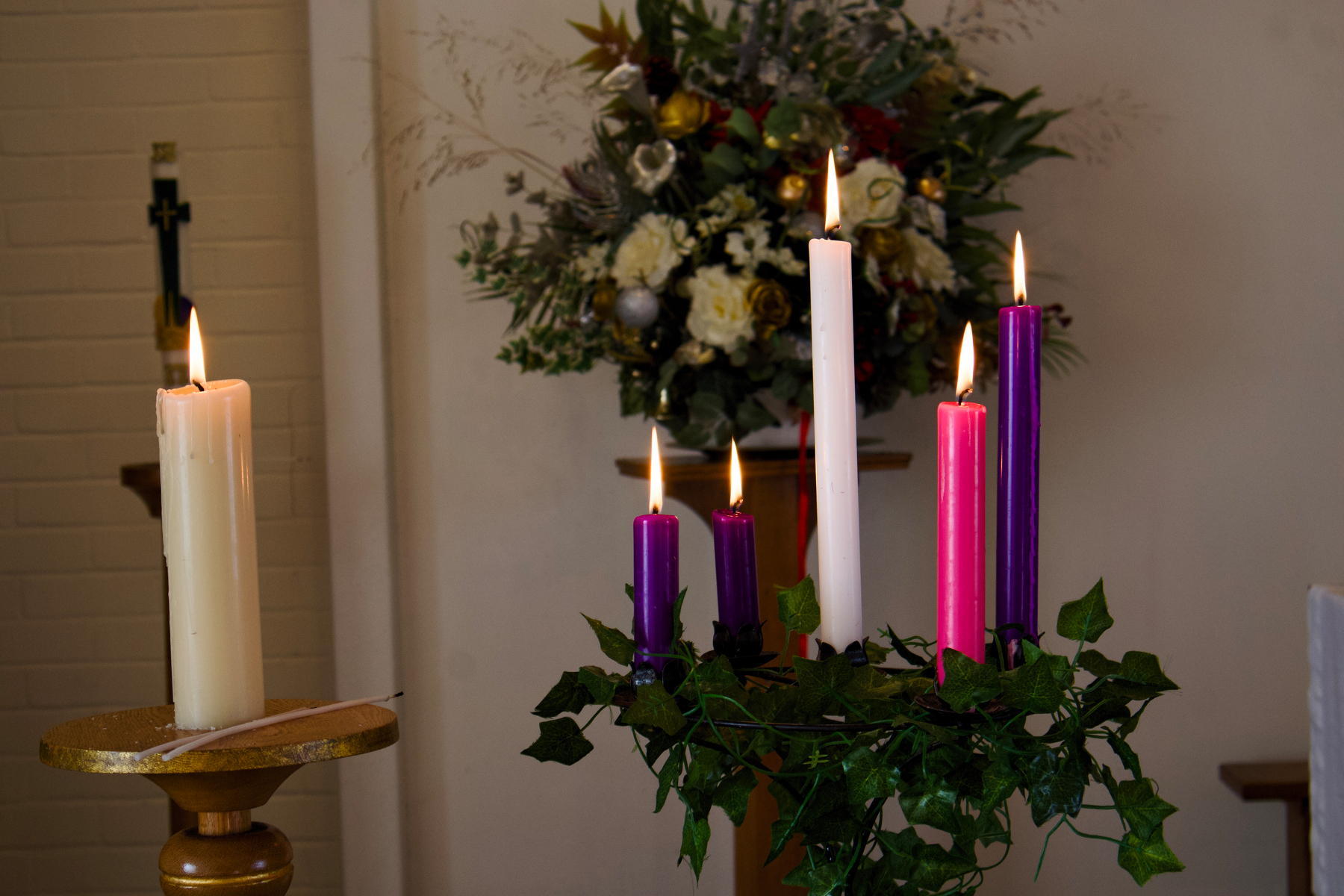 A set of five lit candles, including purple, pink, and white, is surrounded by greenery in a decorative setting with a floral arrangement in the background.