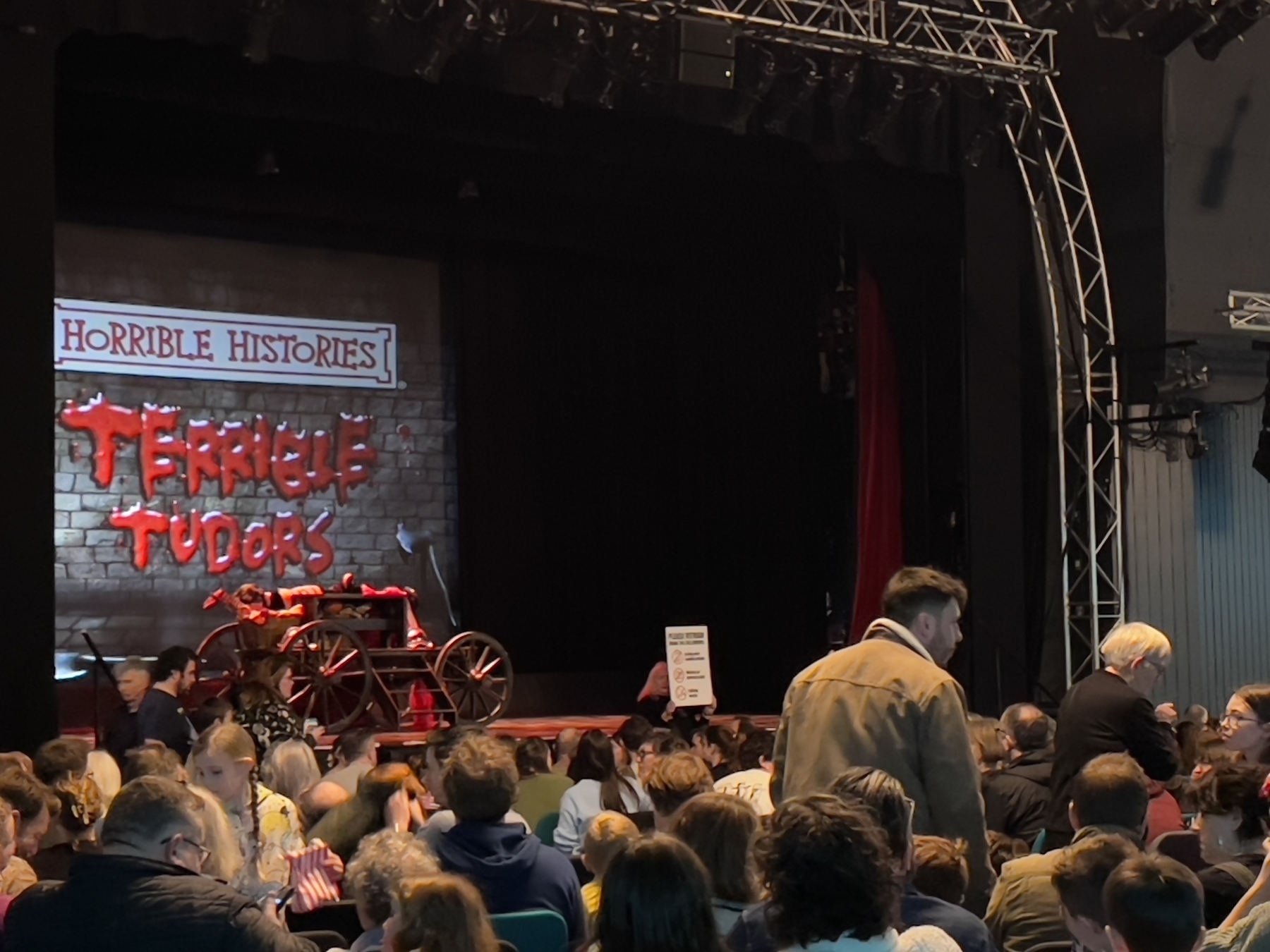 A crowd of people is gathered in front of a stage set for a "Horrible Histories: Terrible Tudors" performance.