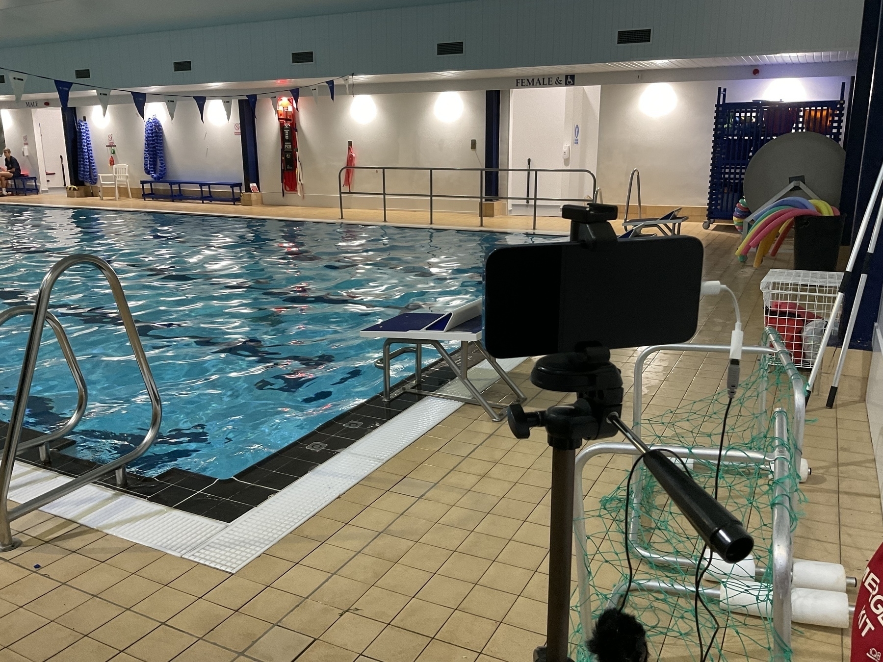 A smartphone mounted on a tripod stands near an indoor swimming pool with people swimming.