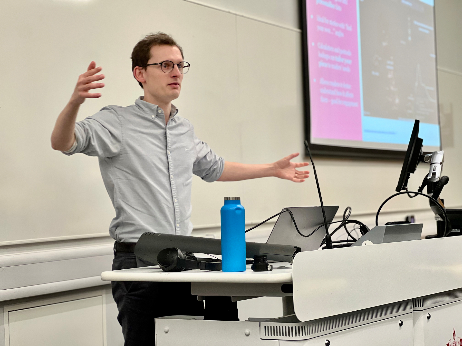 Ashley Kirk is giving a presentation in a conference room, gesturing with open arms beside a projector screen and a table with a water bottle.