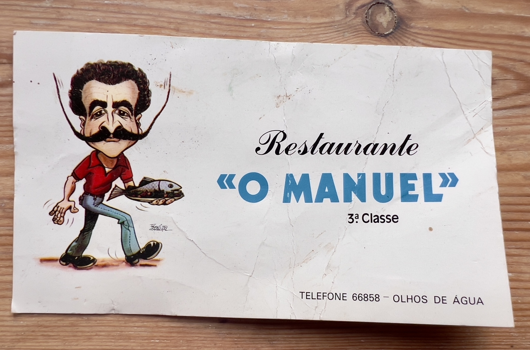 A cartoon man with a large mustache and red shirt is carrying a fish on a restaurant card labeled "Restaurante 'O Manuel'."