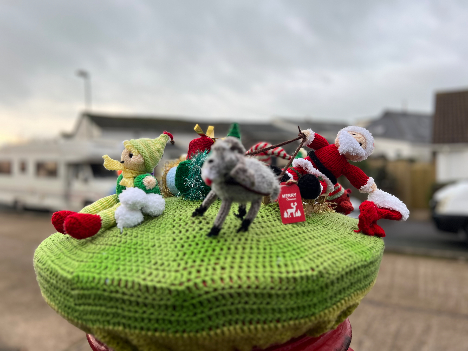 A festive display featuring knitted figures, including Santa Claus, an elf, and a reindeer, is arranged on a green knit-covered surface outdoors.