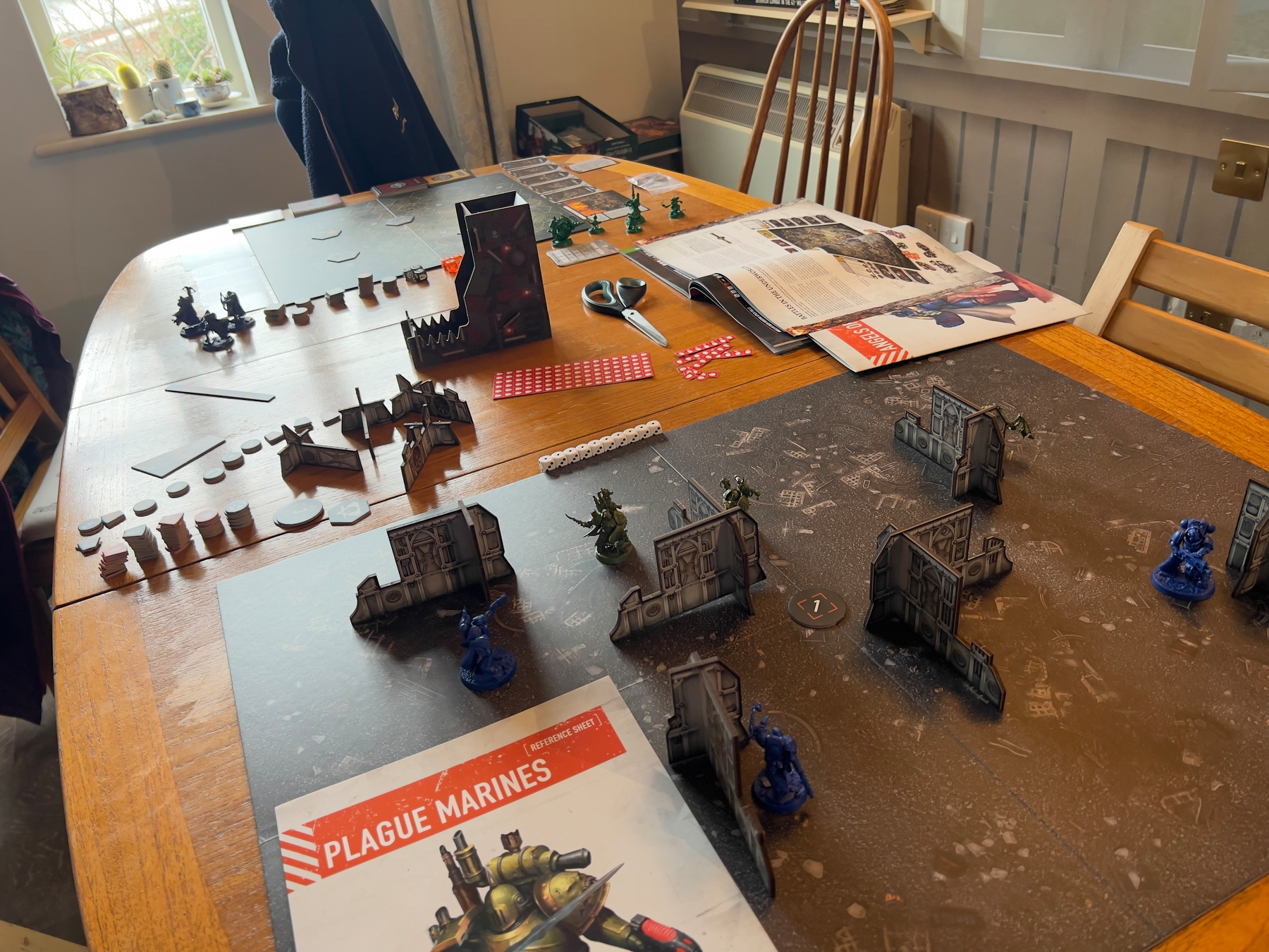 A tabletop wargame setup is displayed with miniatures, terrain pieces, rulebooks, and dice on a wooden table.