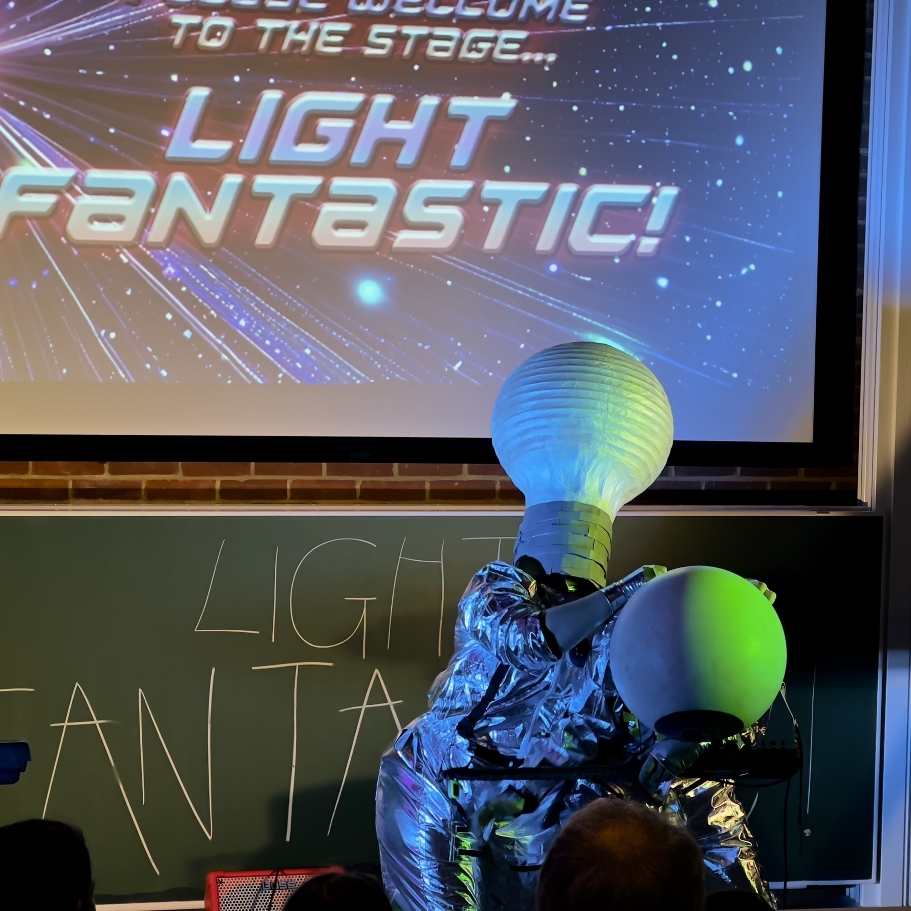 Two people in shiny metallic costumes with large spheres on their heads perform on stage under a sign that says "Welcome to the Stage: Light Fantastic!" while a chalkboard displays similar text.