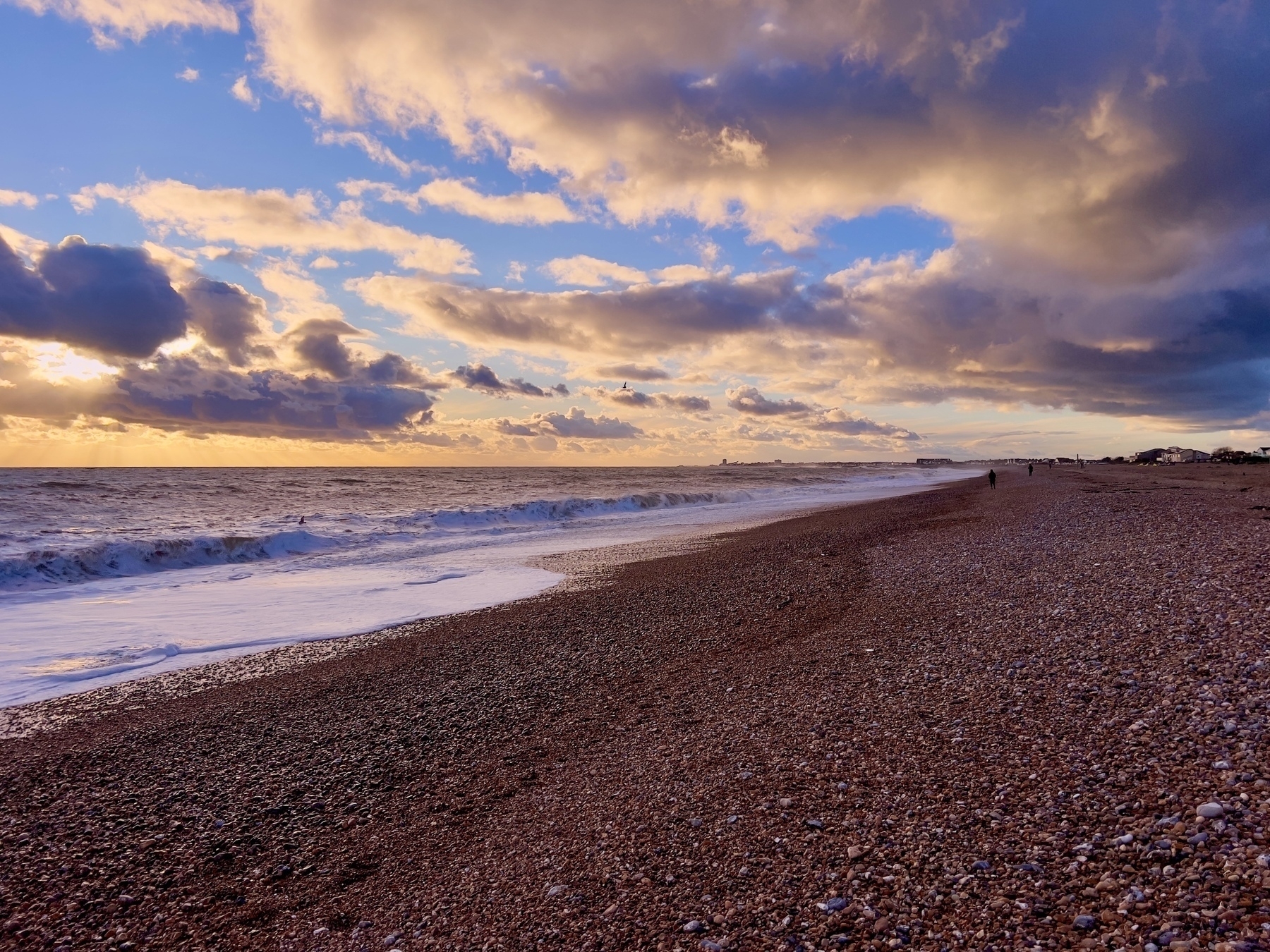 A pebbled beach stretches out to meet the ocean under a dramatic sky filled with colorful clouds during sunset. Worthing pier is visible on the horizon. 