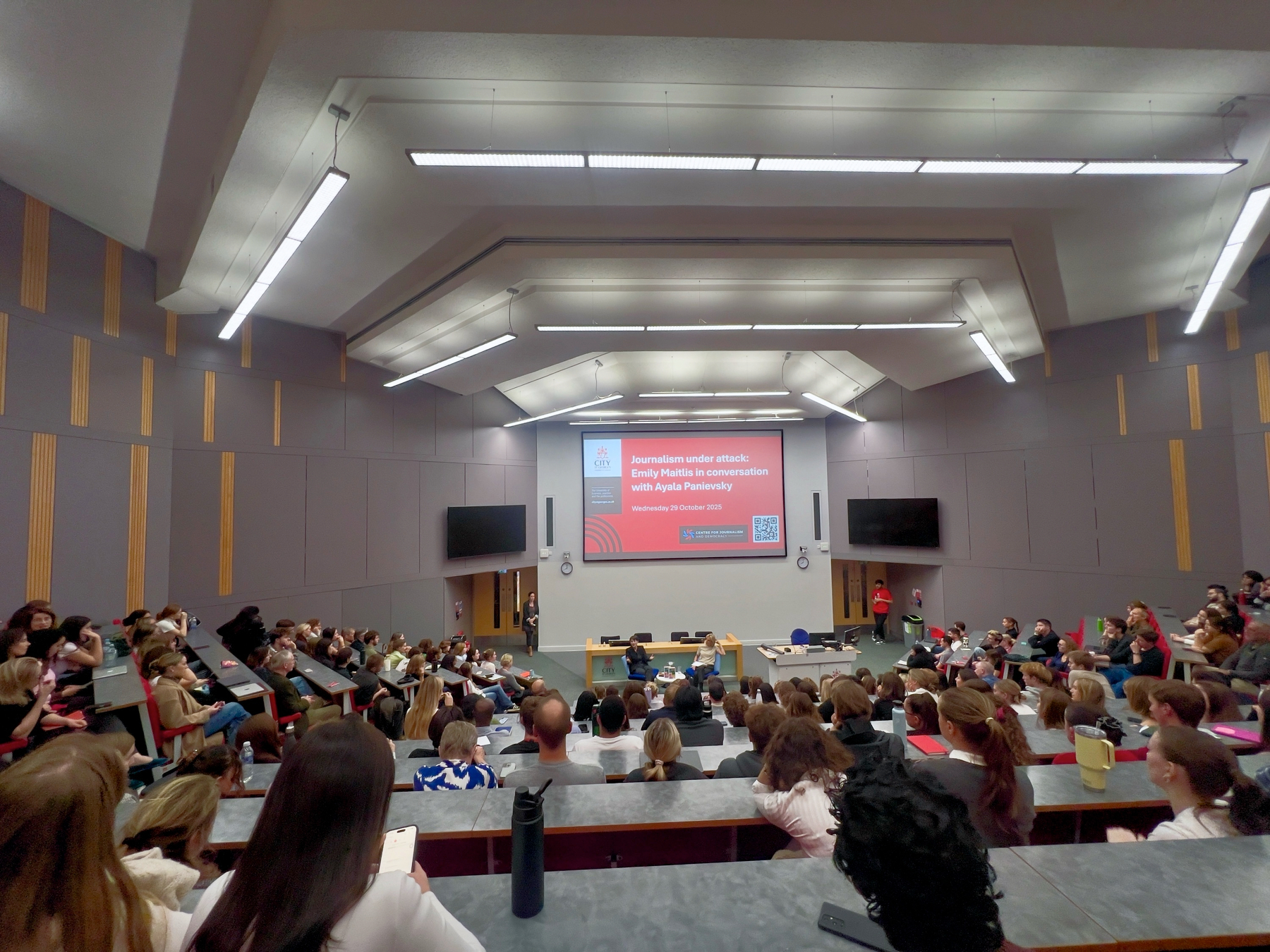 A large lecture hall is filled with people attending a talk between Emily Maitlis and Ayala Panievsky, with a screen displaying the title “Journalism under attack”.