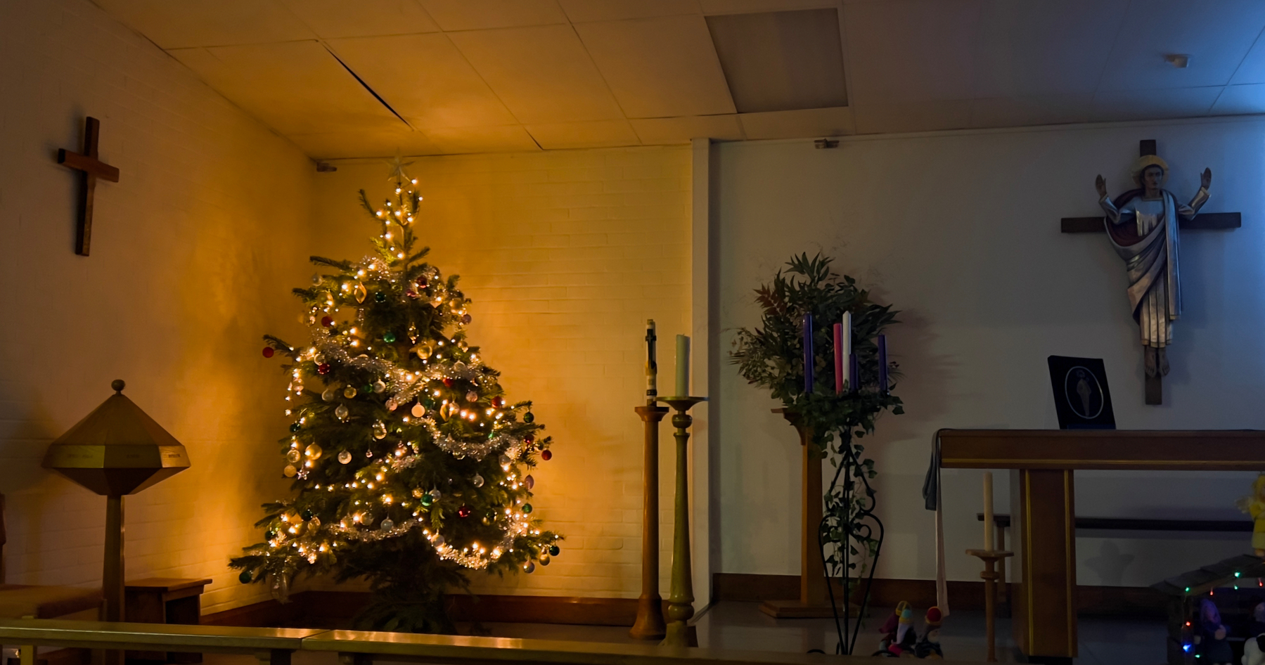 A room is decorated with a lit Christmas tree and Advent wreath in a church setting, featuring two wall crosses.