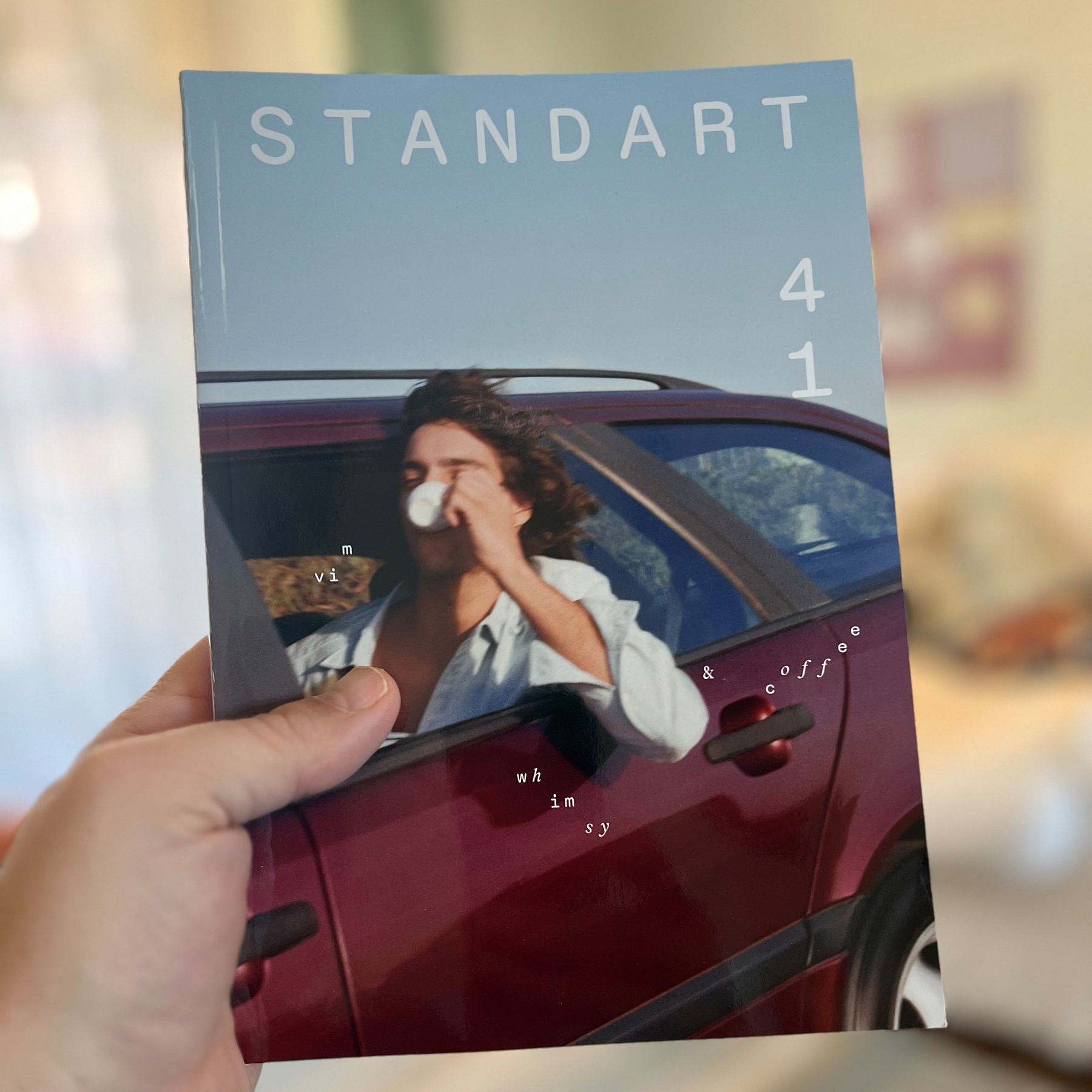 A hand holds a magazine titled STANDART featuring a person drinking coffee while leaning out of a car window.
