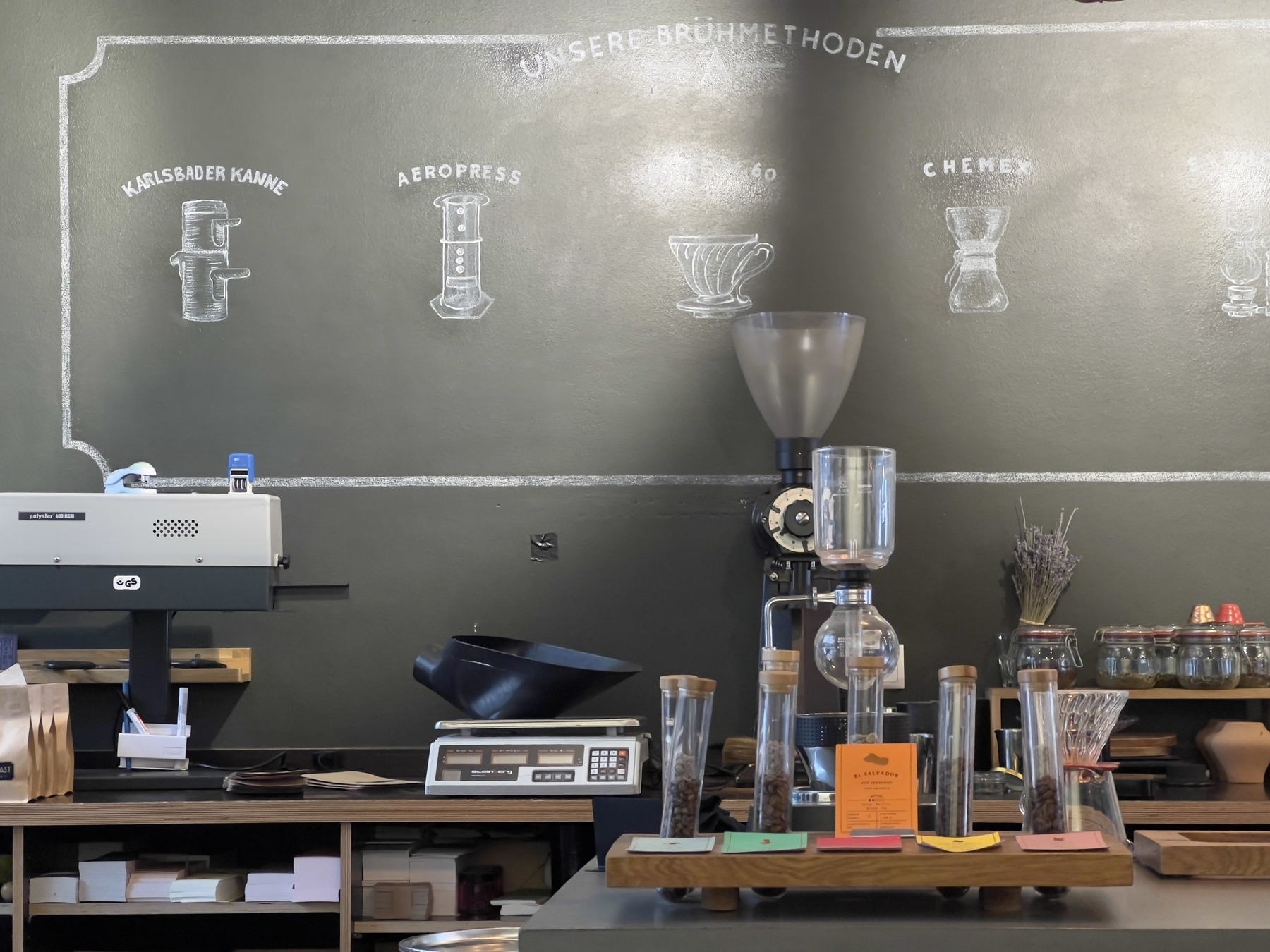A coffee shop counter features various brewing equipment with illustrations of different brewing methods on the wall.
