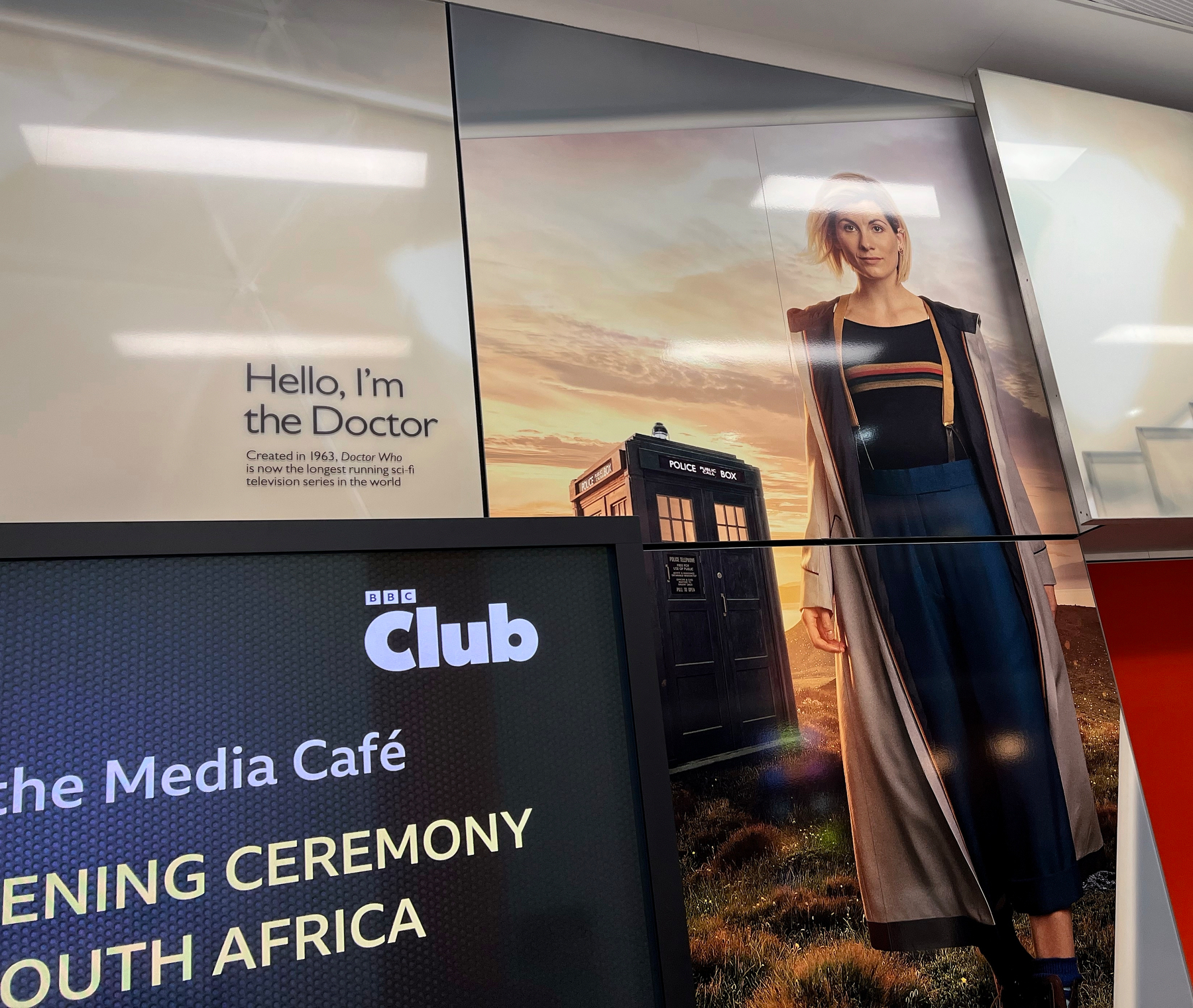 A poster features a character from Doctor Who standing next to the TARDIS, with a digital screen showing an event announcement in the foreground.