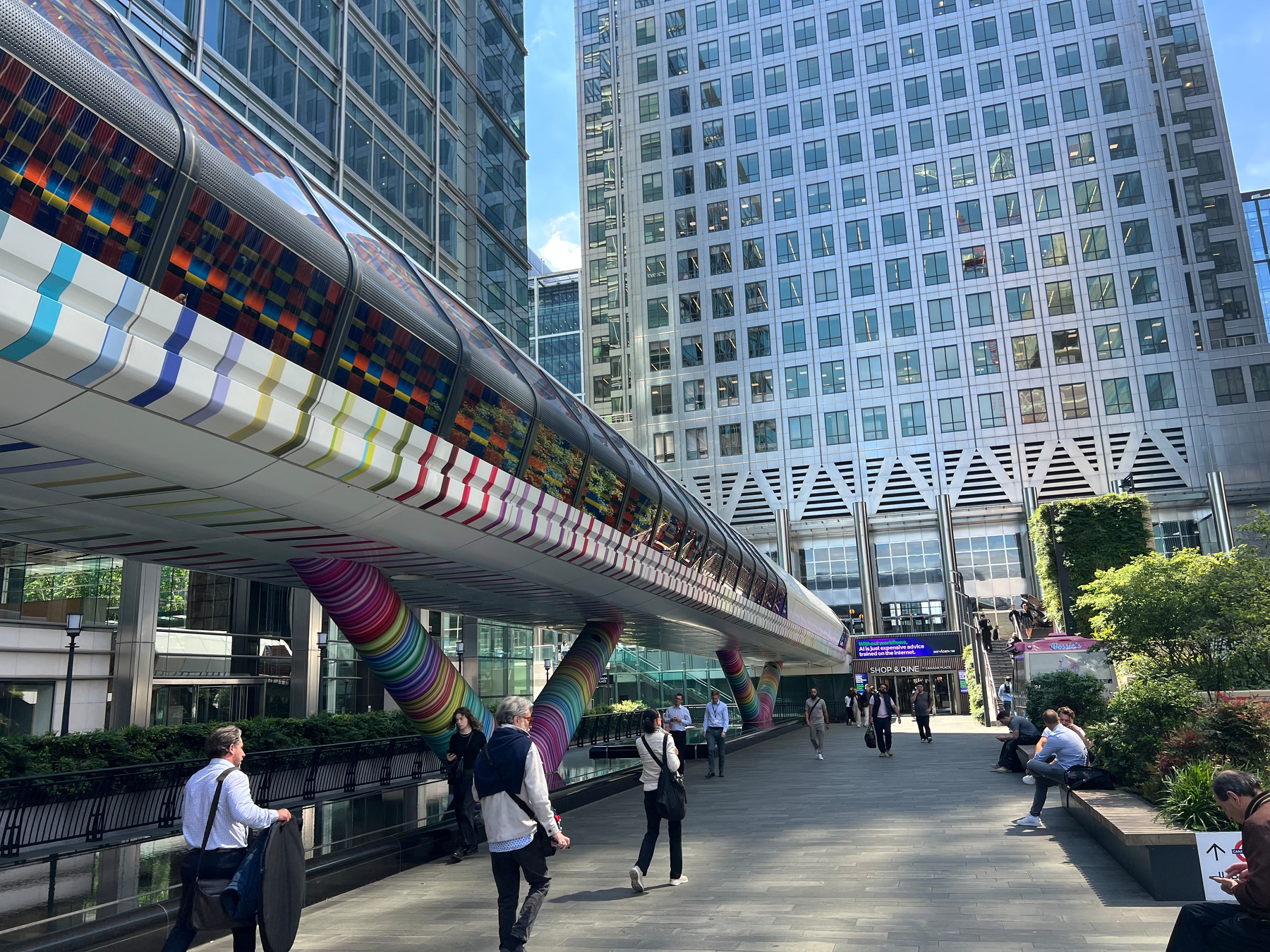 A vibrant, colorful pedestrian bridge connects modern office buildings in a bustling urban area, with people walking and sitting nearby.