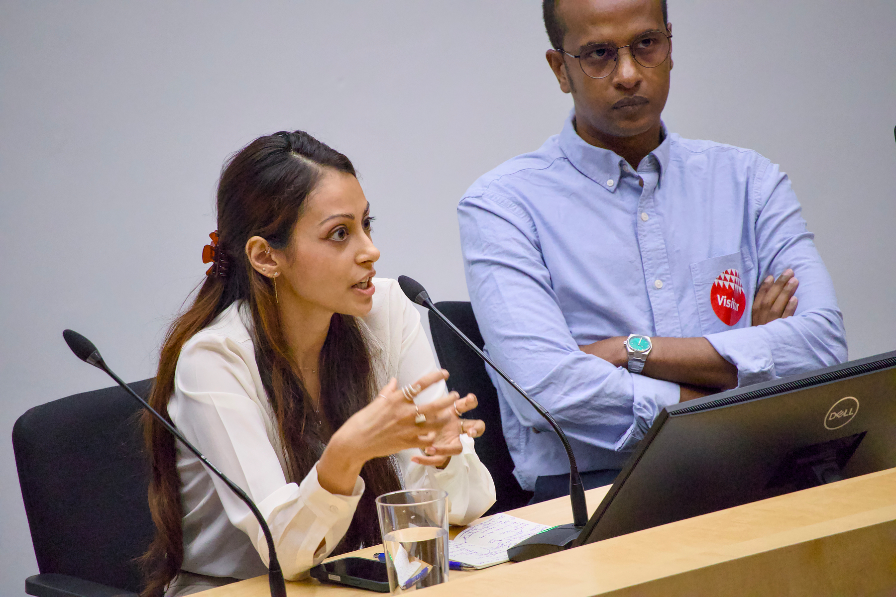 A woman is speaking into a microphone while a man sits beside her with his arms crossed.