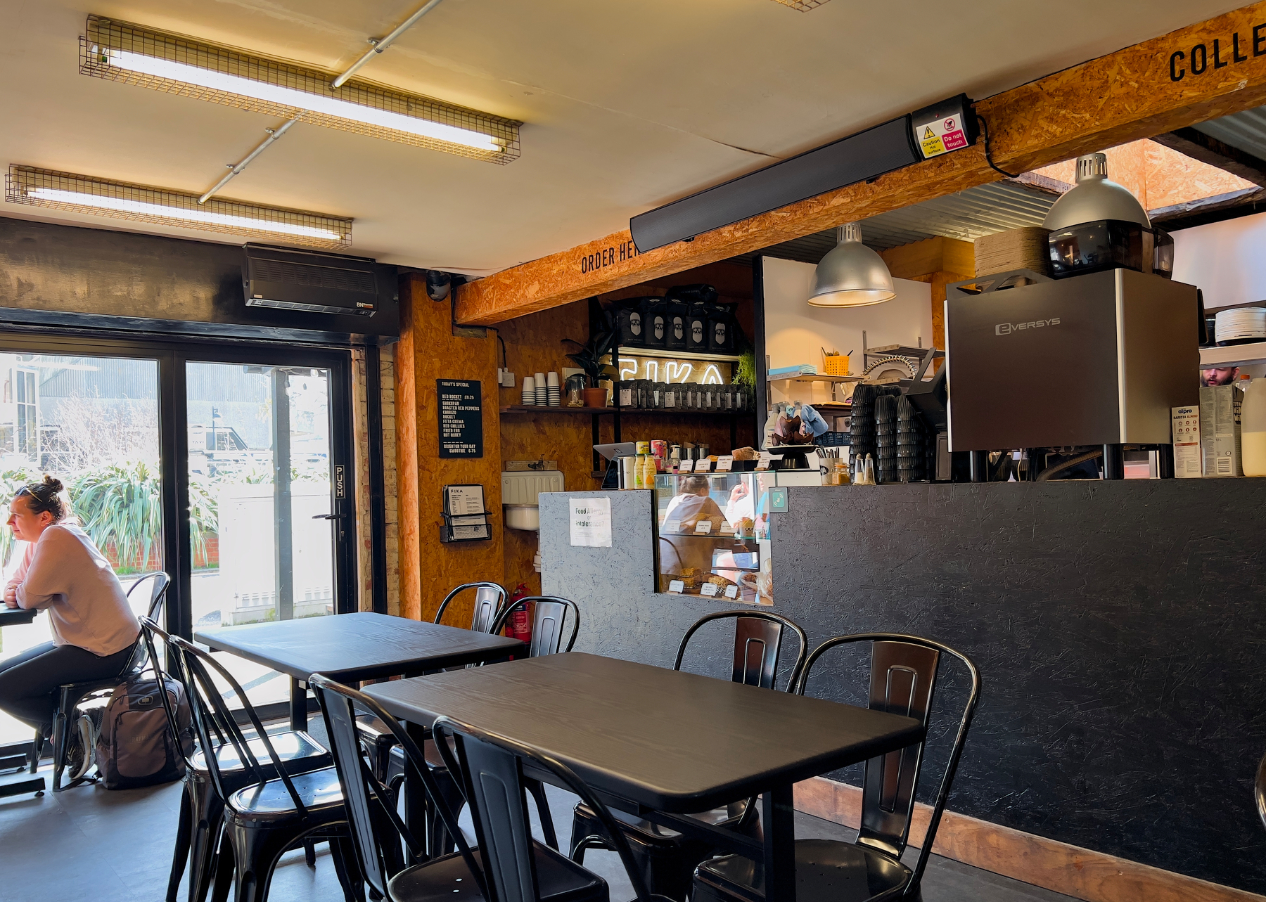 A cozy coffee shop interior features black tables and chairs, a counter with a coffee machine, and a person seated by the window.