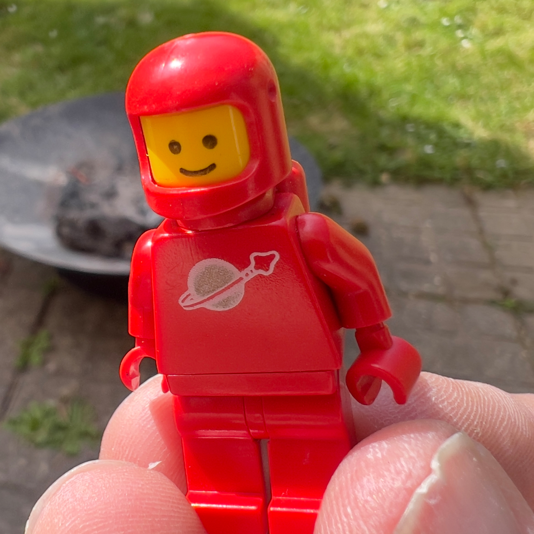 A red LEGO astronaut minifigure is being held outdoors with a blurred background of grass and a fire pit.
