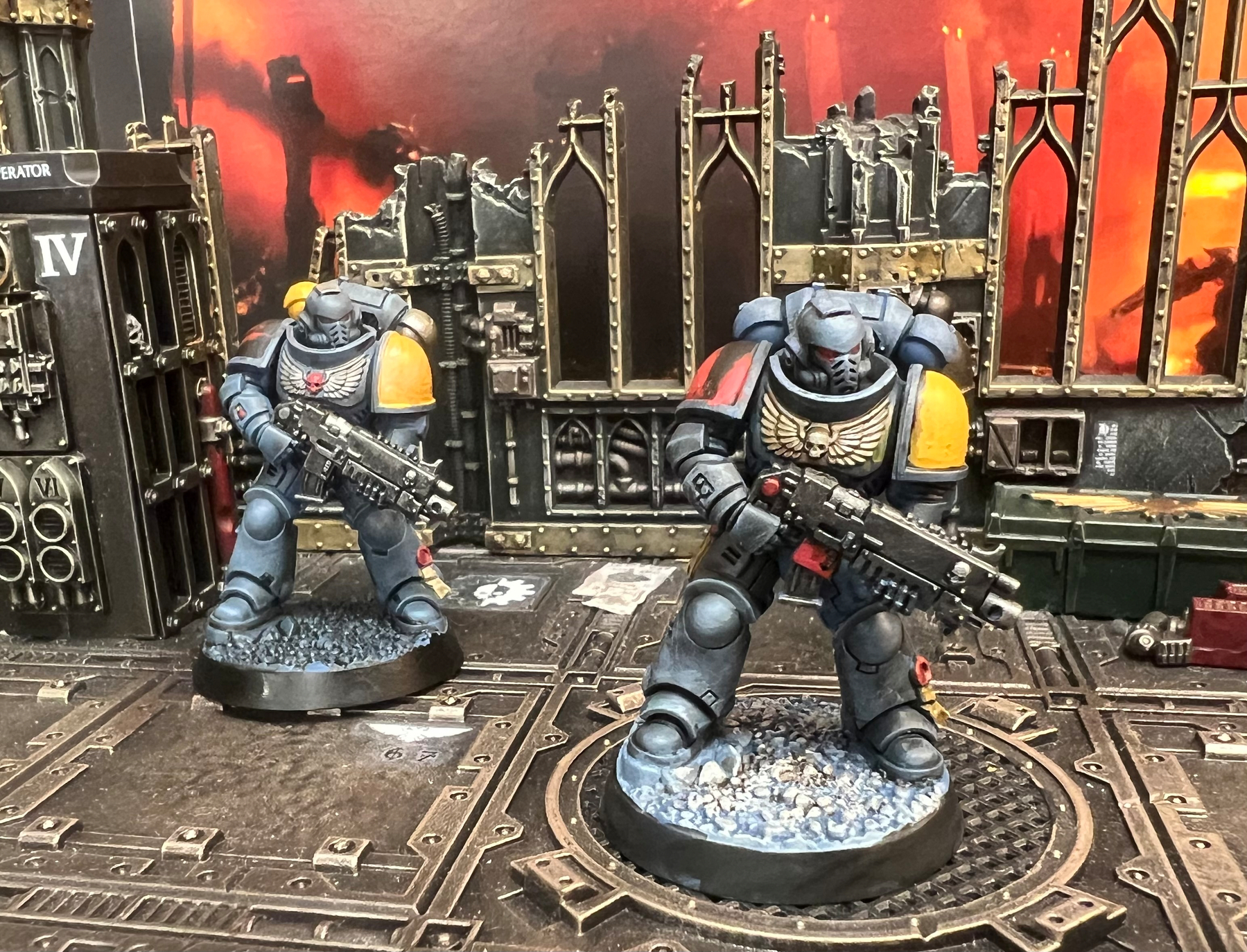 Two intricately painted Warhammer 40k Space Marine miniatures are positioned on a detailed, dystopian battlefield terrain with ruined structures and fiery background.