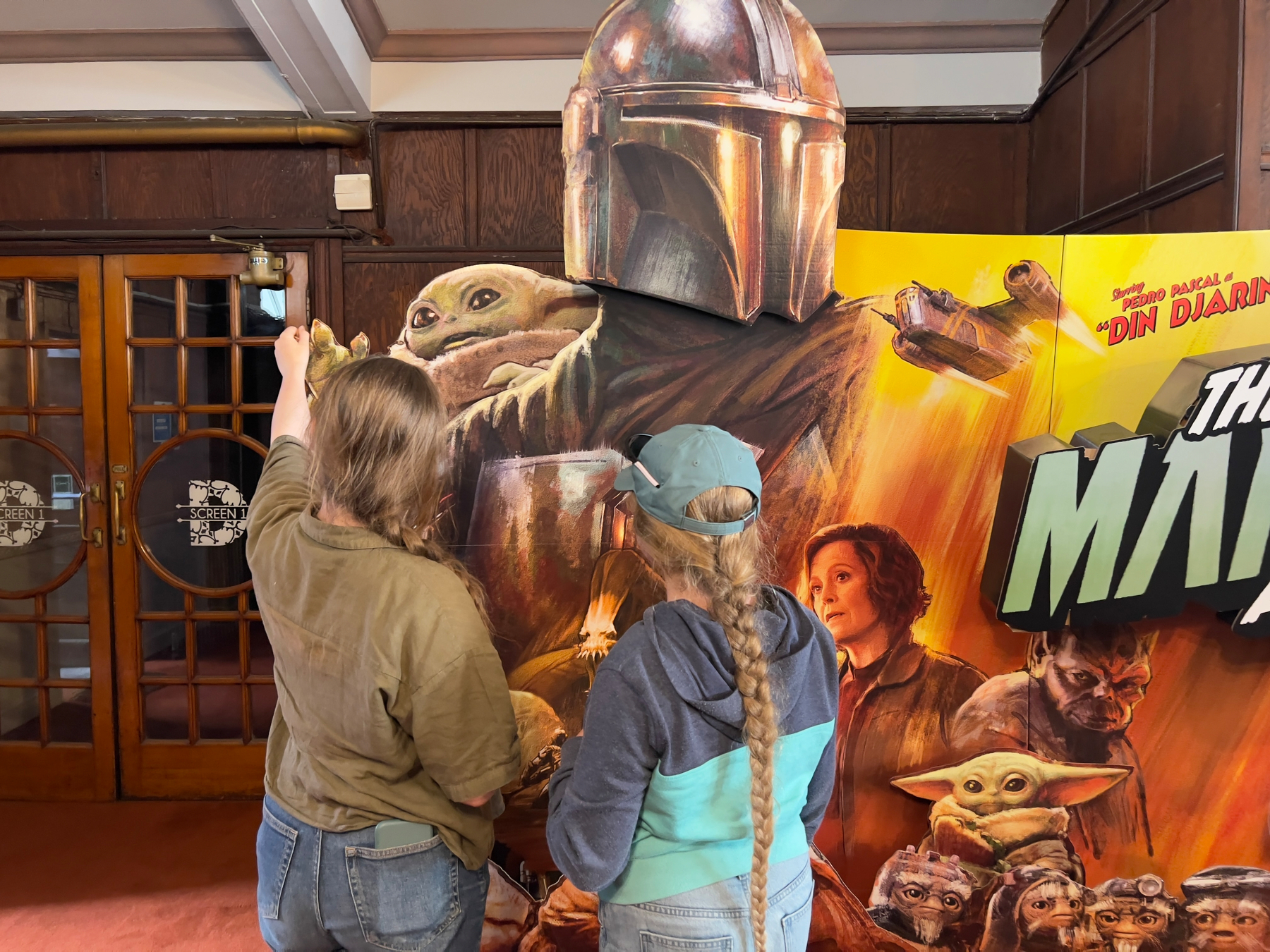 Two people stand in front of a The Mandalorian cardboard display, with one person playfully placing a small figure near it.