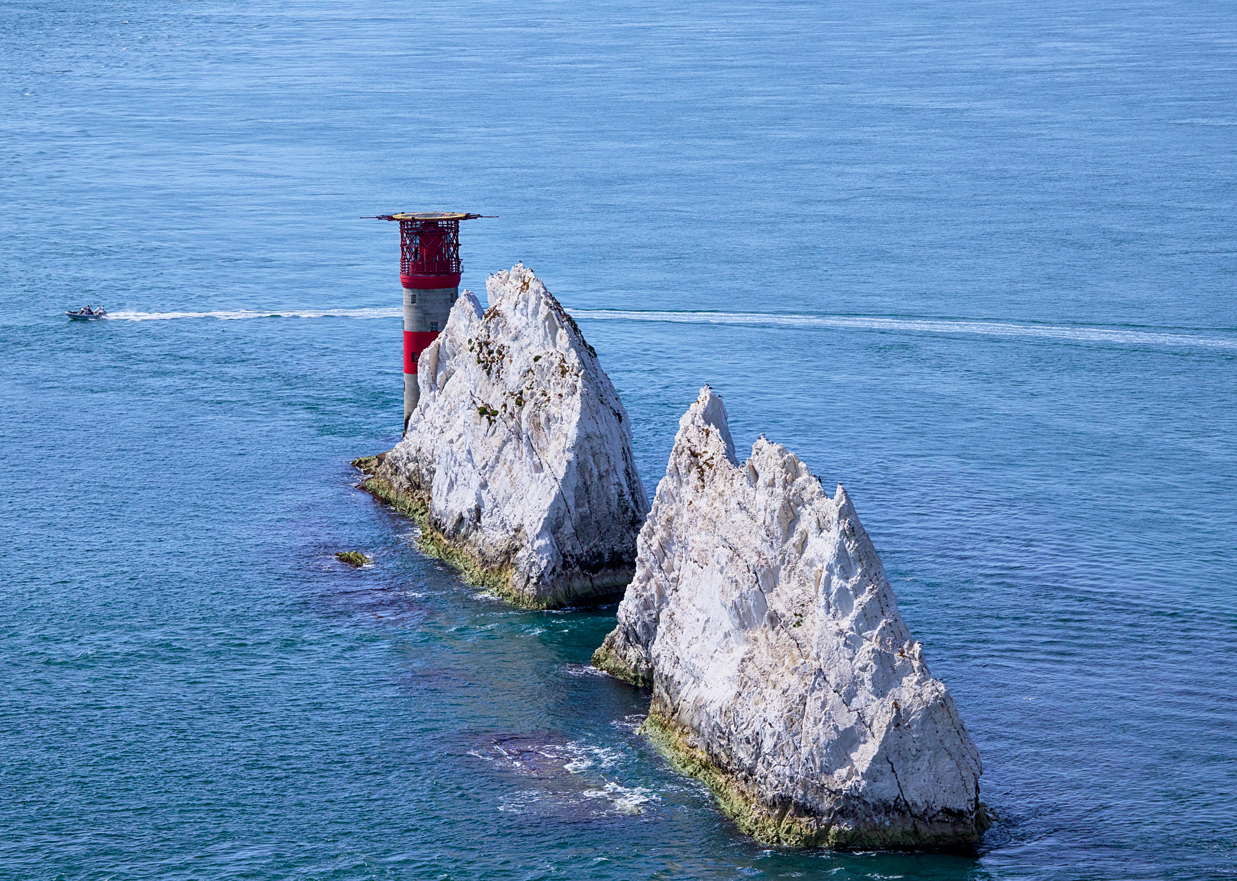A red and white lighthouse stands among two large white rock formations jutting out of the blue sea, with a small boat creating a wake in the water nearby.