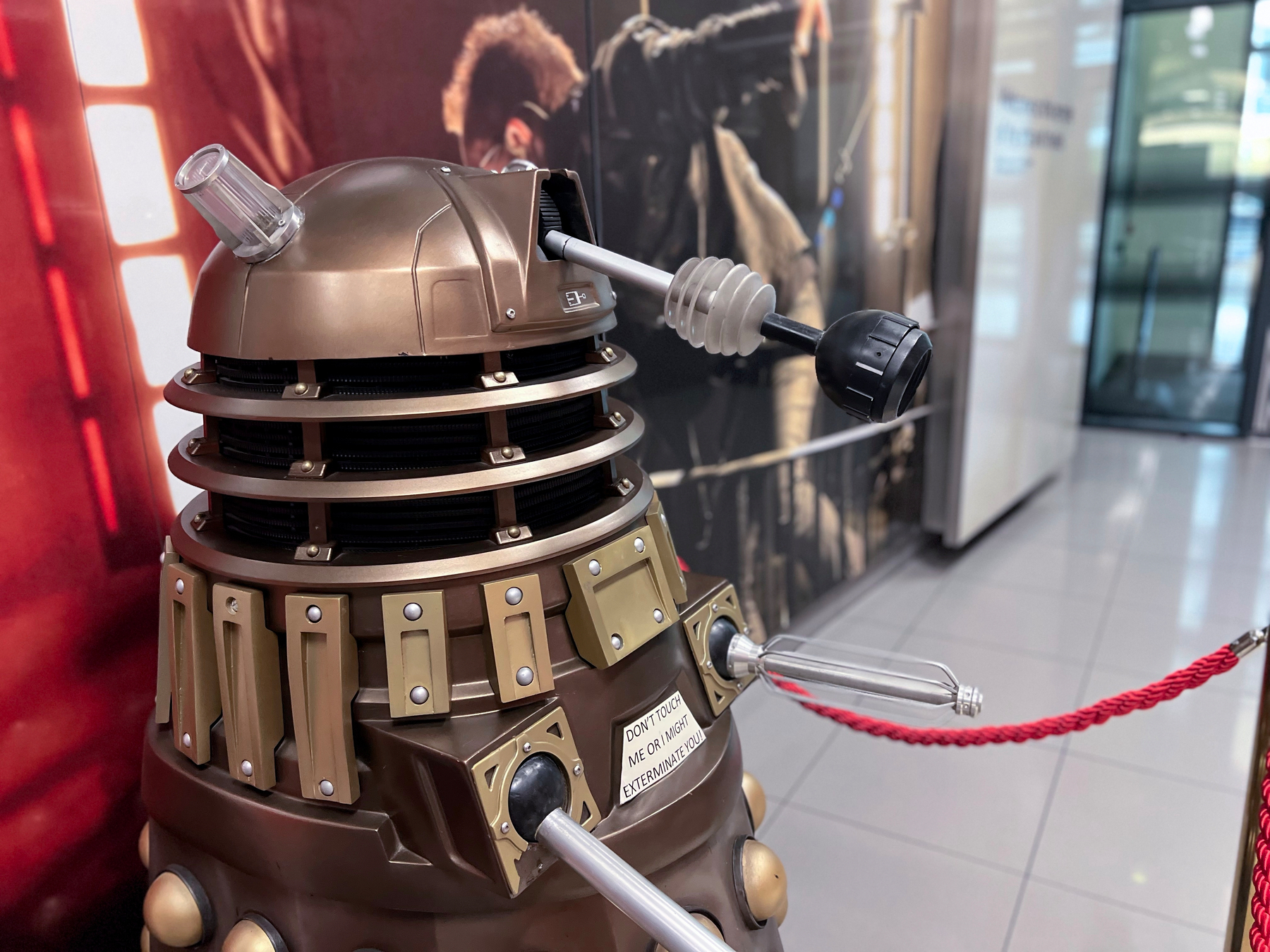 A bronze Dalek model from Doctor Who is displayed indoors, surrounded by a red rope barrier.