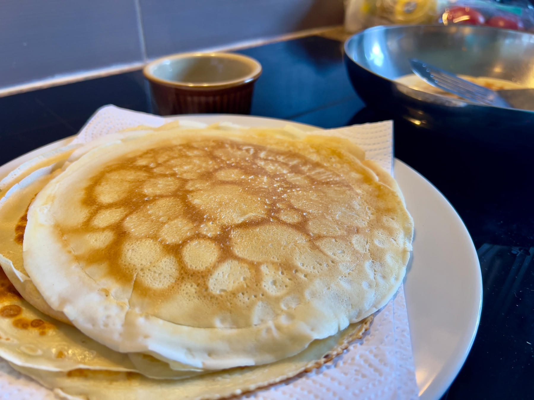 A plate of freshly cooked pancakes rests on paper towels beside a pan and a small ramekin.