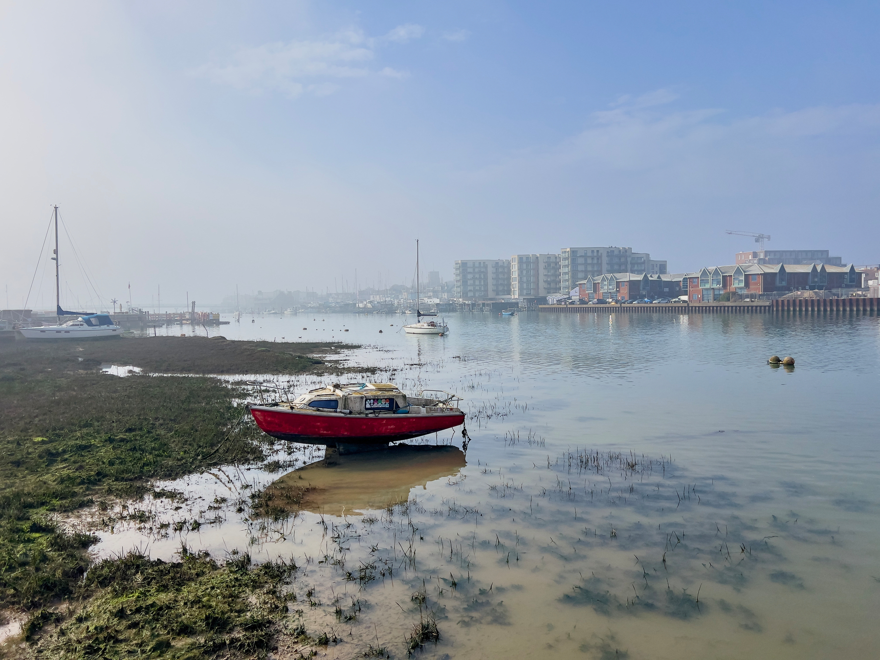 A small red boat is moored on the calm waters of a marina, with misty skies, distant boats, and modern buildings in the background.