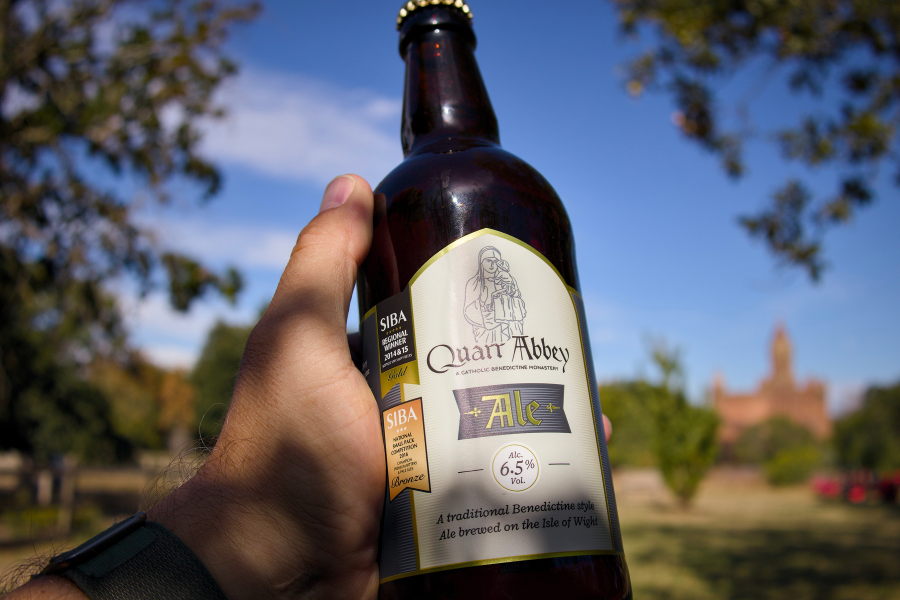 A hand is holding a bottle of Quarr Abbey Ale with a park and the Abbey visible in the background.