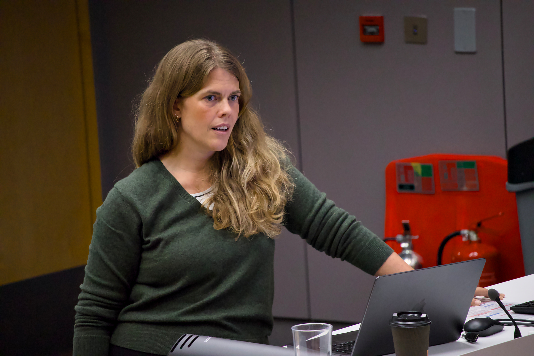 A woman with long blonde hair wearing a green sweater is standing at a desk with a laptop, a cup, and a glass of water, appearing to be engaged in a discussion or presentation.