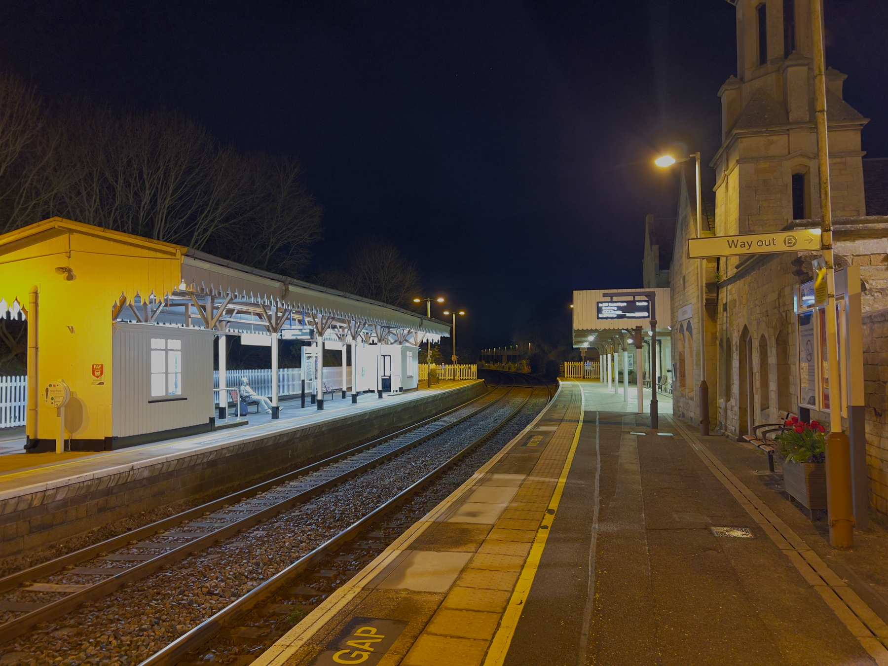 A quiet, illuminated train station platform is seen at night with signs directing to the exit.