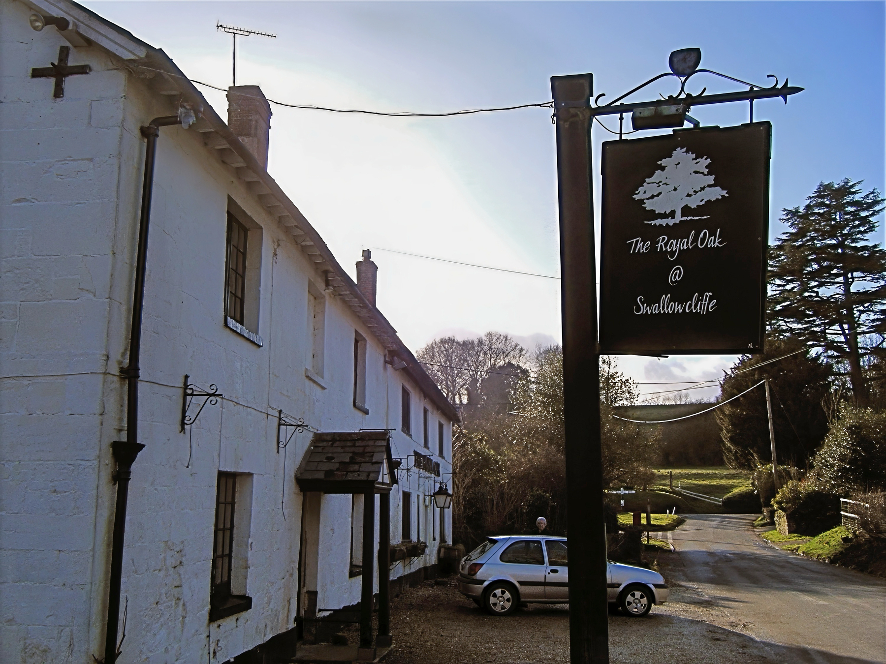 A rural scene features a white building with a sign for The Royal Oak @ Swallowcliffe and a parked car in a countryside setting.