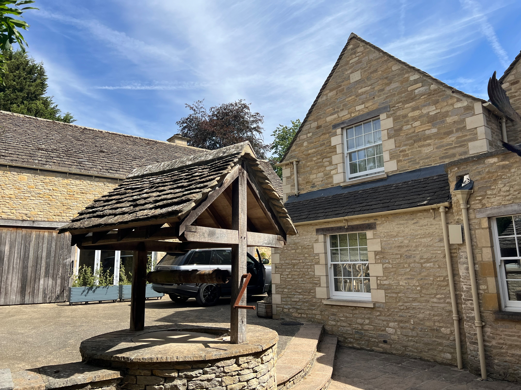 A stone well with a wooden roof stands in front of a traditional stone house under a bright blue sky.