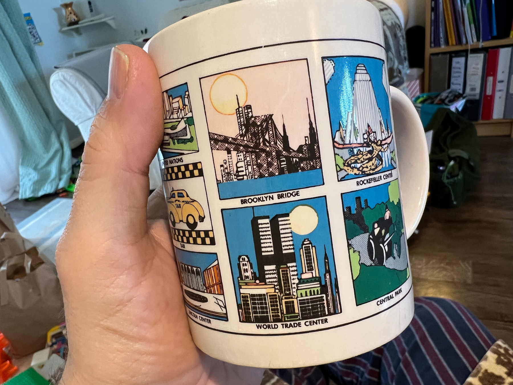 A hand holds a mug featuring colorful illustrated panels of New York City landmarks such as the Brooklyn Bridge and the World Trade Center.