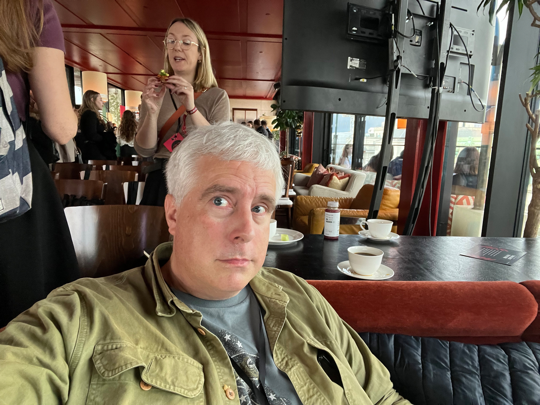 A man with gray hair is sitting at a table in a café or restaurant with cups and plates, while a woman standing behind him appears to be speaking or eating.