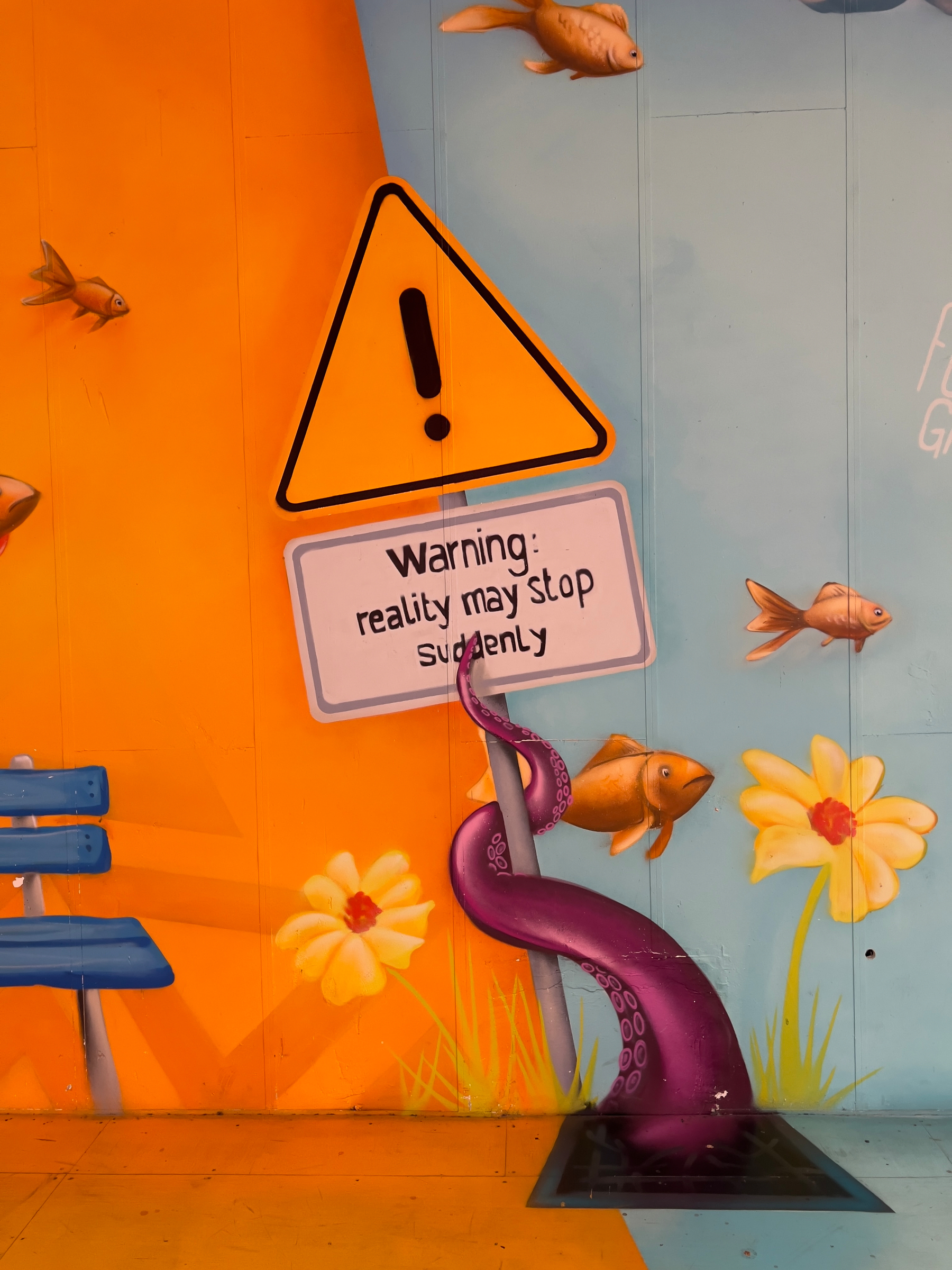 A whimsical mural features a warning sign with reality may stop suddenly, surrounded by fish, flowers, a purple tentacle, and a blue bench.