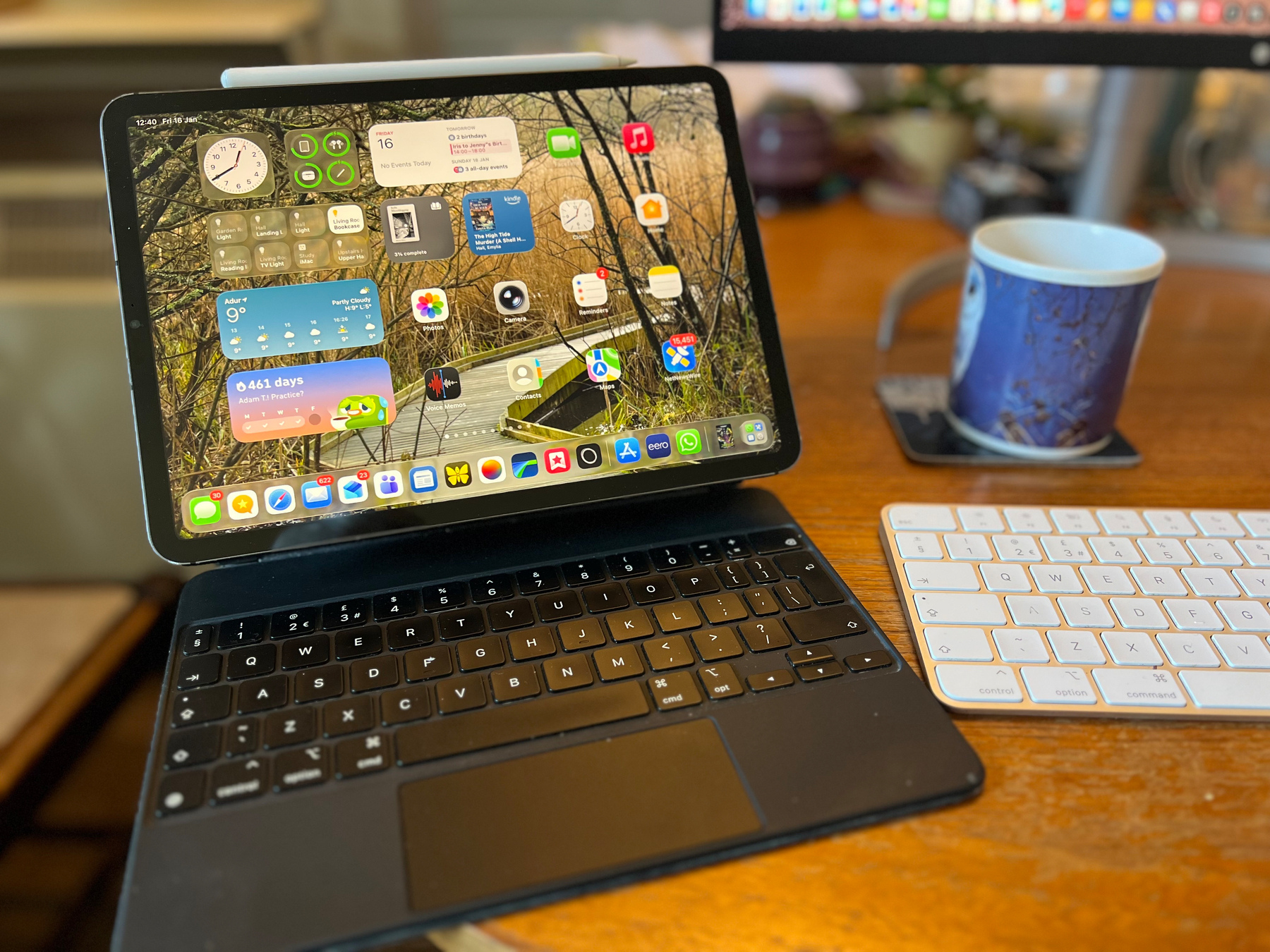 An iPad with a keyboard case displaying a home screen full of apps and widgets on a wooden desk next to a wireless keyboard and a coffee mug.