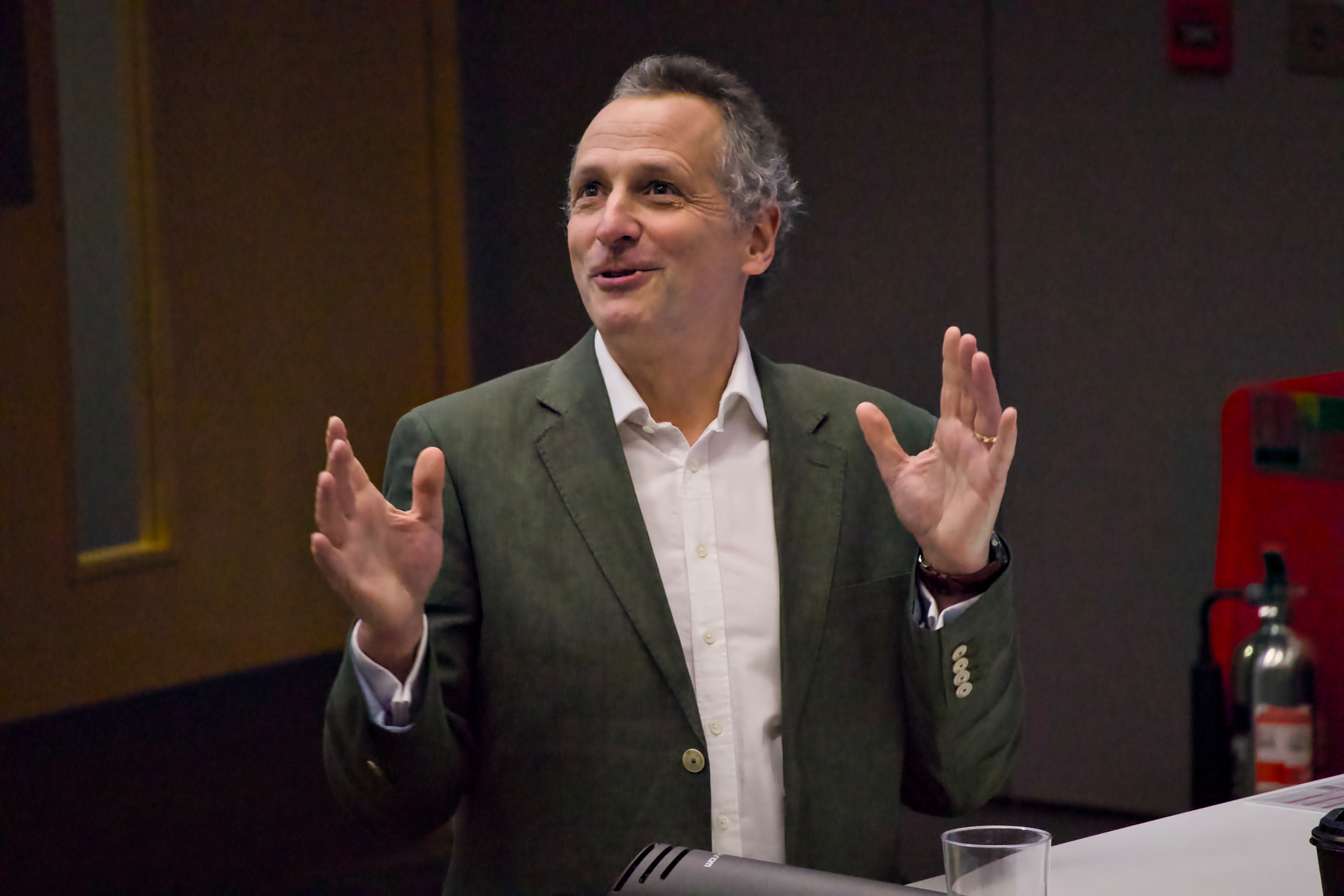A man wearing a green blazer and white shirt is speaking with expressive hand gestures in an indoor setting.