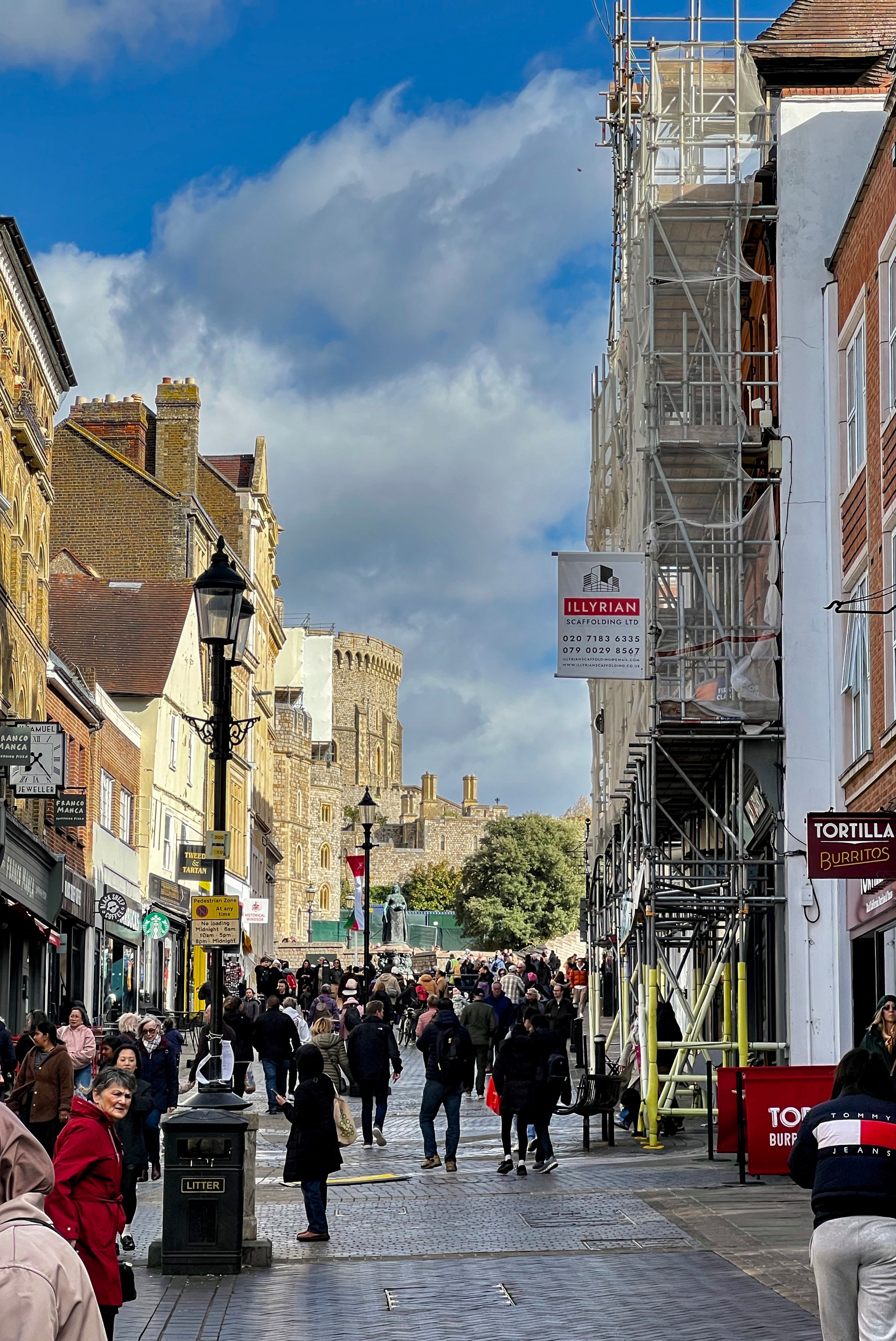 A busy pedestrian street filled with people walking toward a historic castle under a partly cloudy sky.