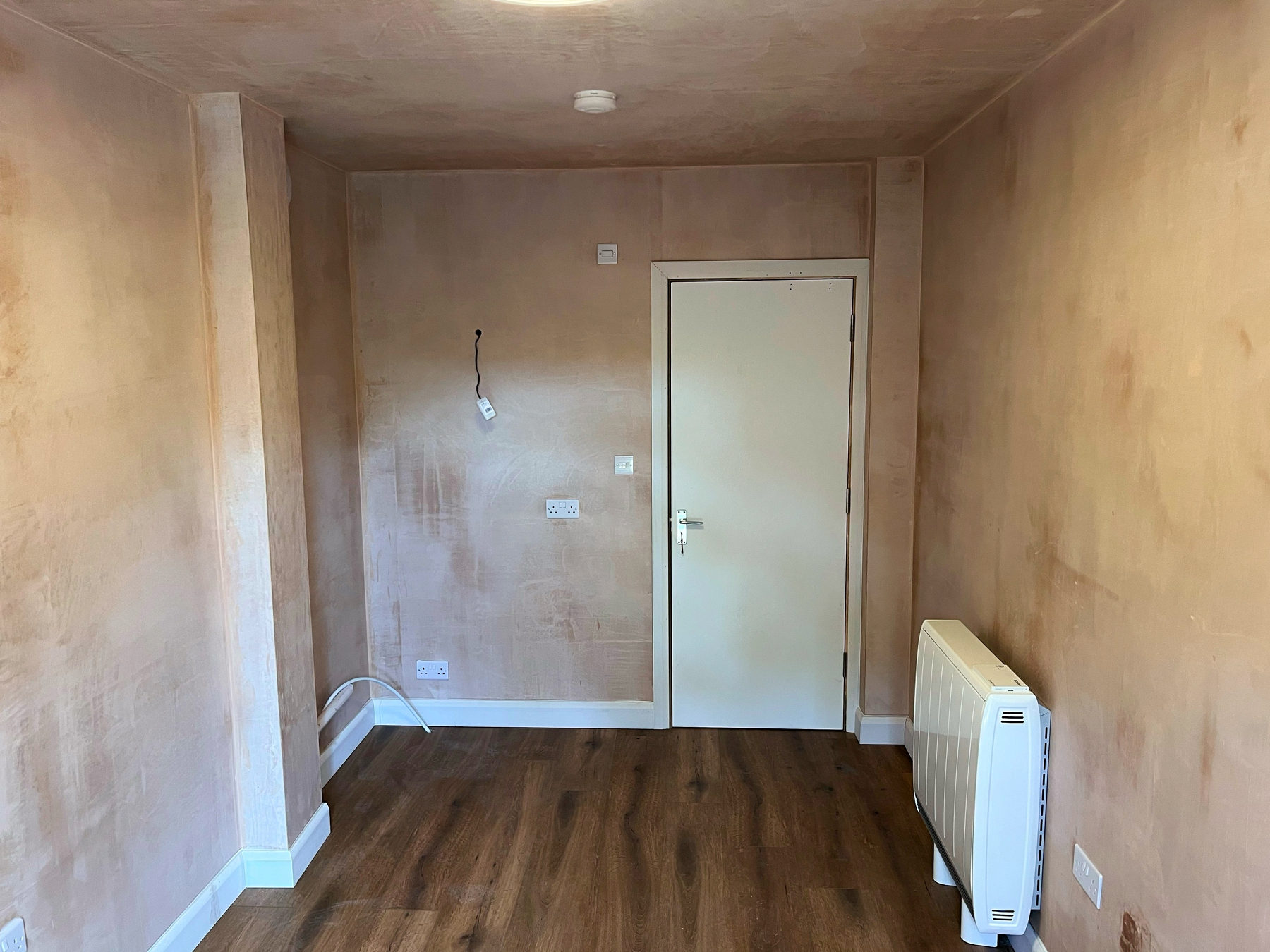 A small, newly plastered room with a wooden floor, radiator, and a white door.