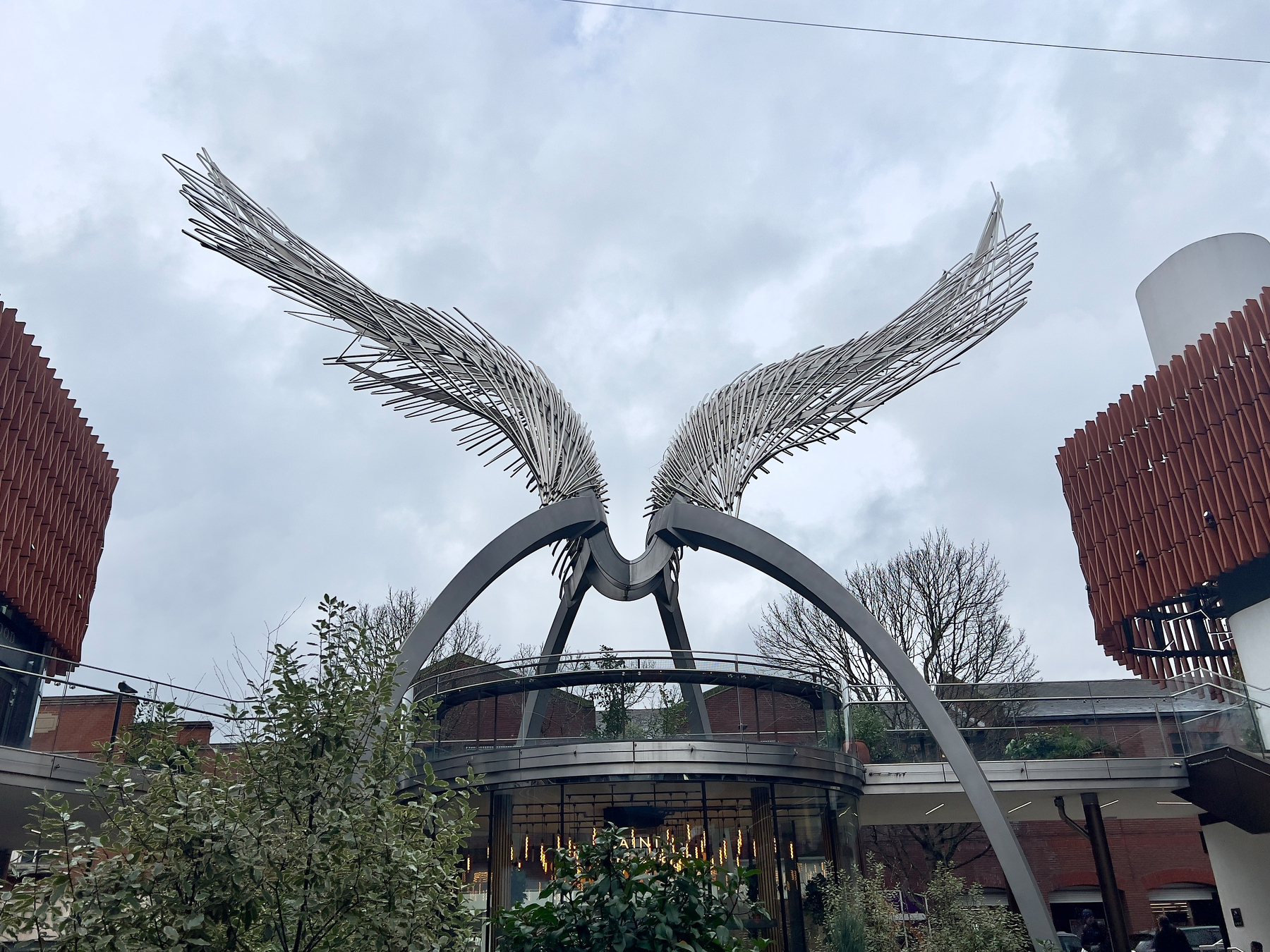 A large, intricate metal sculpture resembling wings stands prominently in an outdoor public space, surrounded by modern architectural elements and greenery.