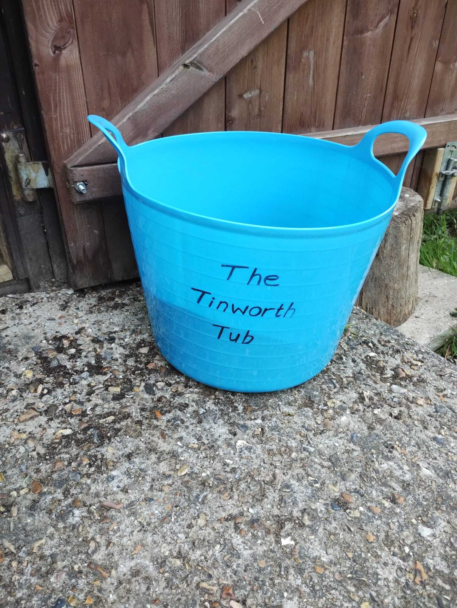 A blue plastic tub labeled The Tinworth Tub is placed on a textured concrete surface near a wooden fence.