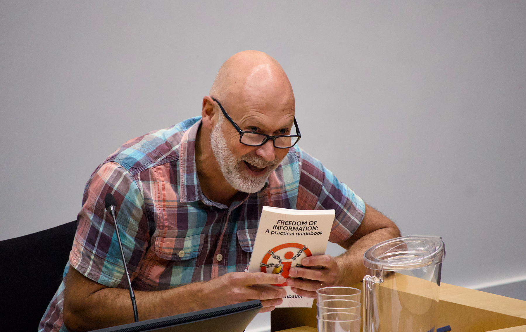 A bald man with glasses wearing a colorful plaid shirt is smiling while holding a book titled FREEDOM OF INFORMATION: A practical guidebook and sitting at a table with a microphone and a glass pitcher.