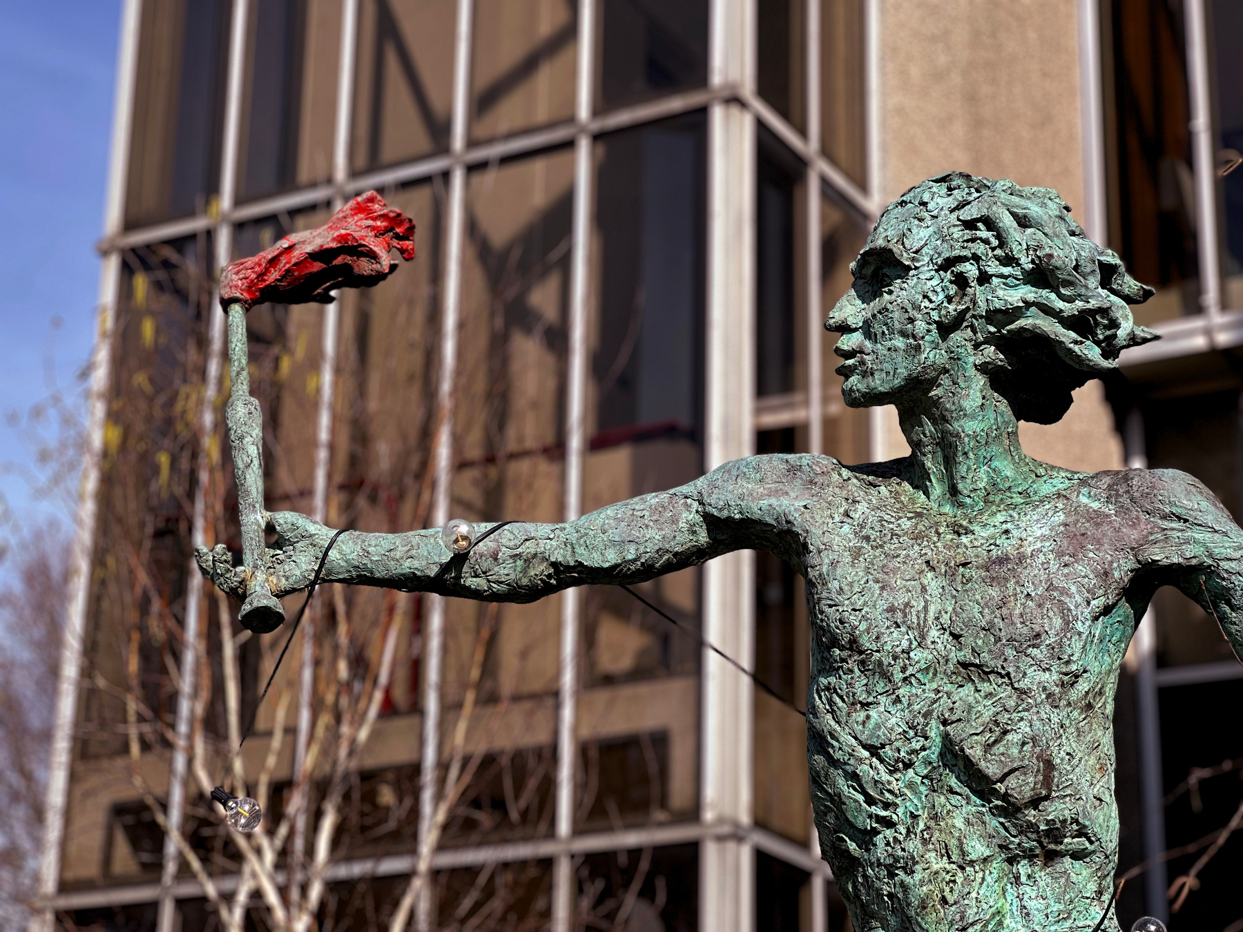 A bronze statue of a person holding a red flower is positioned in front of a building with large windows.
